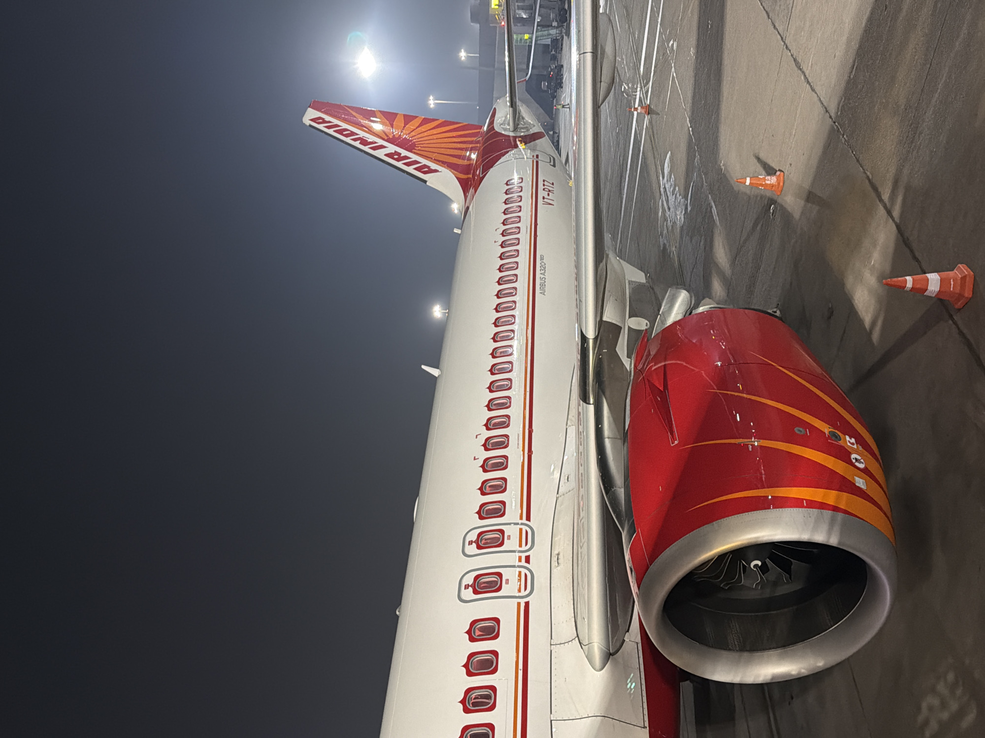 An Air India Airbus A320neo, registration VT-RTZ, is parked on the tarmac at Indira Gandhi International Airport in New Delhi, India. The aircraft's distinctive red and orange livery is visible on the tail and engine nacelles, with flame-like designs on the engines. The airport tarmac is wet, with some puddles and patches of what appears to be ice or snow. Several orange traffic cones are placed on the ground, likely to mark areas or direct movement. In the background, airport lighting and other aircraft can be seen, indicating it is nighttime. The sky is dark and overcast. No people are prominently featured in the foreground or midground. The image captures a detailed close-up of the aircraft's wing, engine, and tail section.