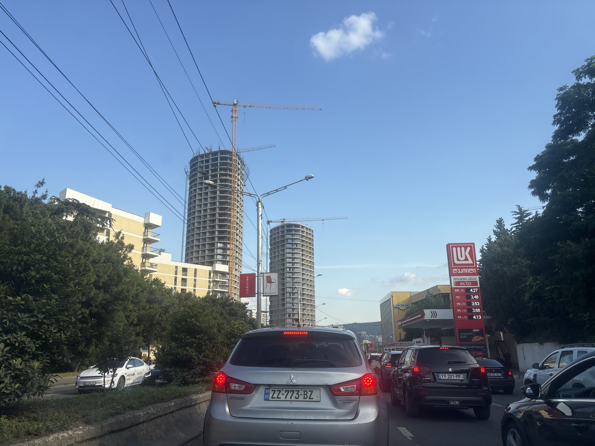 The image captures a street scene in Tbilisi, Georgia, characterized by heavy traffic congestion under a clear blue sky with scattered clouds. In the foreground, the rear of a silver Mitsubishi Outlander Sport is visible, its brake lights illuminated. The vehicle's license plate reads "ZZ-773-BZ". To the left, a white sedan with license plate "GE 343-CC" is partially obscured by trees.
The midground is dominated by two large, unfinished cylindrical skyscrapers under construction, with cranes positioned around them. A street lamp is also visible. To the right, a Lukoil gas station with a prominent red and white sign displays fuel prices. Nearby, a building with a sign reading "BRICORAMA" is visible.
The background shows more of the city's architecture and rolling hills in the distance. Overhead, a network of electrical wires spans across the sky. The overall impression is one of urban development and daily city life, with a focus on the ongoing construction projects.
