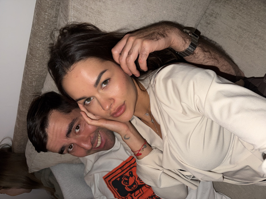 The scene is an indoor, soft-lit setting, depicting two individuals in the foreground seated closely on a light-colored, textured couch. A third individual is partially visible and out of focus in the upper left background. The specific location within the city of Tel-Aviv, Israel, cannot be determined from the visual content, which depicts a private residence or a relaxed social space.
In the foreground, a female with dark hair and green-hued eyes, estimated to be in her 20s or 30s, is positioned closer to the camera. She wears a light beige or cream-colored top with a V-neck and a draped, possibly wrap-style front. Accessories include a delicate silver-toned necklace with small reflective elements and a red string bracelet with small silver-toned charms on her left wrist. A faint crescent moon symbol is visible on the inside of her left wrist. Her left hand is raised, with fingers lightly touching the side of her face near the cheekbone. She looks directly at the camera with a neutral to soft expression.
Behind and slightly to her left is a male, also estimated to be in his 20s or 30s, with dark hair and eyes. He is smiling broadly, looking directly at the camera. He wears a white t-shirt featuring a prominent orange graphic on the left chest depicting a stylized character with large ears. His right arm is wrapped around the female, indicating close interaction. His left forearm, visibly hairy, displays a dark-colored metallic link watch. The overall impression is a posed photograph in a casual, intimate setting.