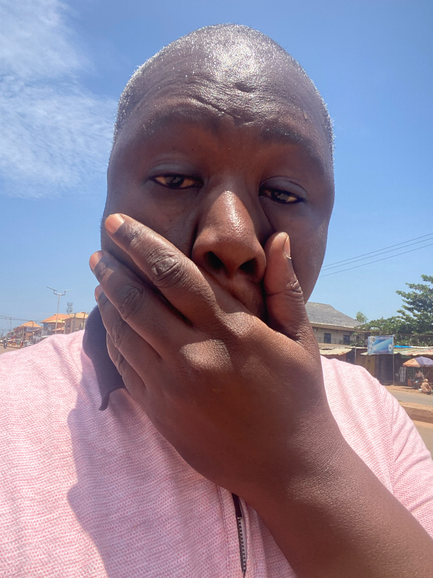A close-up, selfie-style shot shows a person's face from the nose up, with their right hand covering their mouth. The person has dark skin and is wearing a light pink, textured collared shirt. Their forehead and the visible part of their face appear sweaty, with a sheen reflecting the bright sunlight. The background is an outdoor scene under a clear blue sky with scattered clouds. In the distance, buildings with orange roofs are visible along a street lined with shops and market stalls, with people and vehicles present. The overall impression is a sunny day in an urban environment, possibly a market area in Sabon Tasha, Nigeria, with the subject captured mid-gesture.