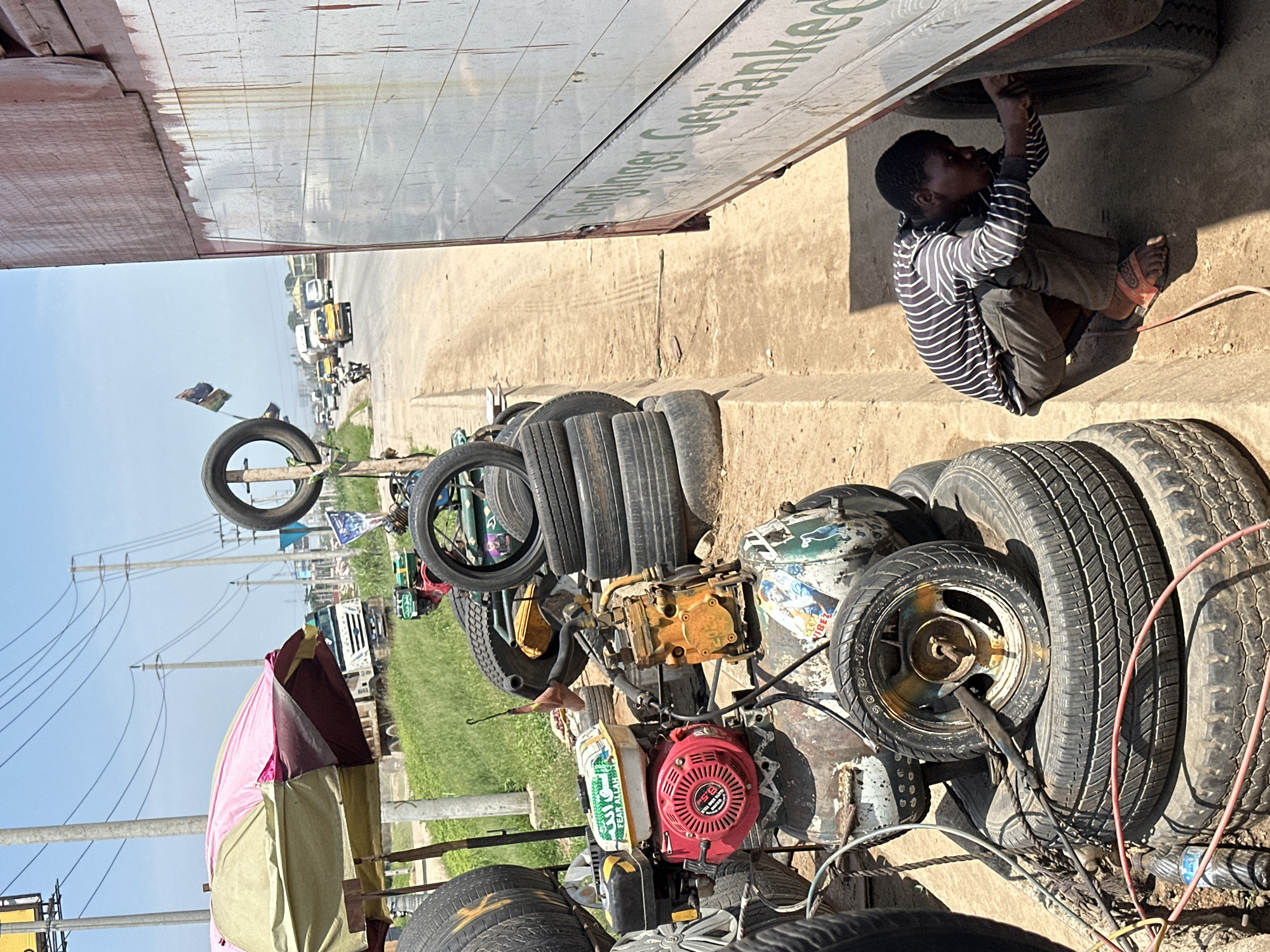 The image depicts an outdoor tire repair shop located on the side of a road in Magboro, Nigeria. A young boy, wearing a striped long-sleeved shirt and dark pants, is crouched down, working on the tire of a large truck or bus parked on the right side of the frame. The truck's body is partially visible, with lettering that includes "Tenglinger Getränke".
In the foreground and middle ground, numerous used tires are piled high. A red and yellow air compressor unit, powered by a small engine with the inscription "FEAR ALLAH" on it, sits among the tires. Various hoses and electrical cords are scattered around the repair area. An umbrella, a mix of green, pink, and brown, is set up to provide shade.
The background shows a busy road with several vehicles, including cars, trucks, and motorcycles, moving under a bright, clear sky. Utility poles with numerous wires stretch across the landscape. The general environment appears to be a roadside commercial area with a mix of vegetation and infrastructure. The ground is a mix of dirt and concrete.