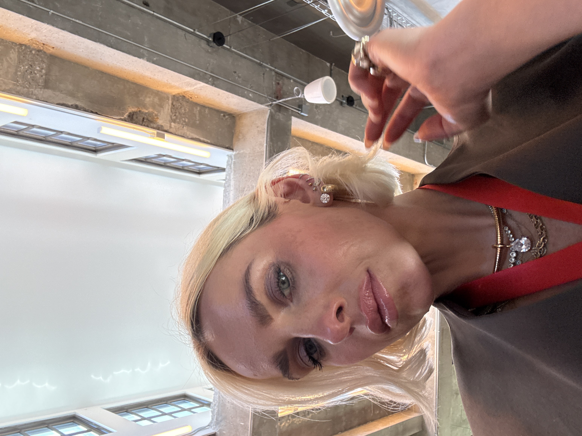 A woman with blonde hair and light skin is shown in an indoor setting. She is wearing earrings and necklaces. Her fingers are visible near her hair. The background includes a concrete ceiling with lighting fixtures and windows. The location appears to be in Los Angeles, United States of America, based on the architectural style and ambient lighting.