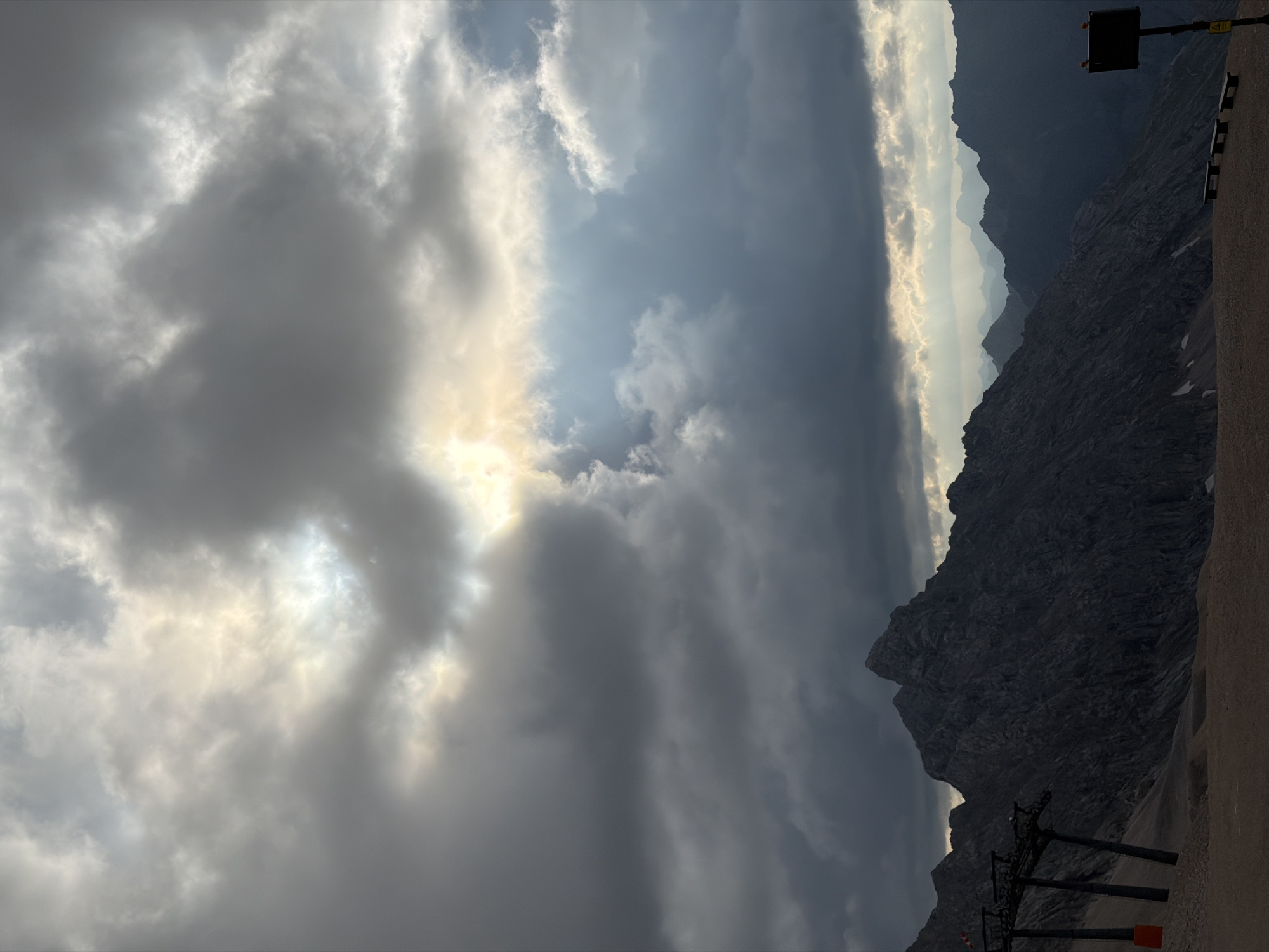 This is a photograph of a mountainous landscape, likely taken from a high vantage point. The sky is filled with dramatic, dark clouds with rays of sunlight breaking through in several places, creating a sense of depth and atmosphere. In the foreground, there are rocky mountain slopes. On the left side of the image, part of a ski lift structure with its supports and cables is visible, suggesting this location might be a ski resort. Further into the background, multiple layers of mountain ranges recede into a hazy distance. The ground in the foreground appears to be a sandy or gravelly surface. The overall mood of the image is somber and majestic due to the lighting and the imposing mountain peaks. This scene is consistent with the summit of the Zugspitze, Germany's highest peak, which is located near Garmisch-Partenkirchen.