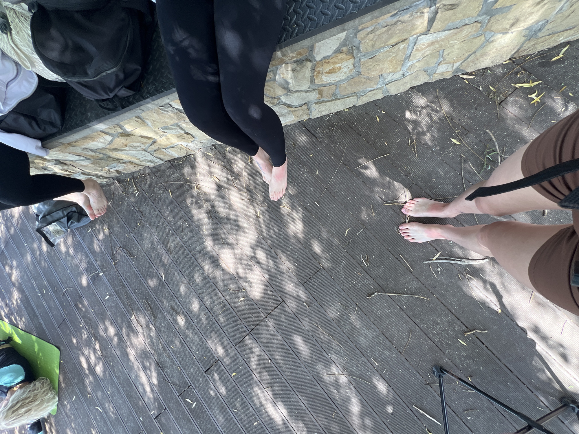 The image shows a group of people sitting and lying on a wooden deck near a stone wall. In the foreground, a person's legs and feet are visible, wearing brown shorts. Further back, two people are sitting on the stone wall, both wearing black leggings. One person is lying on a green mat, facing away from the camera, with blonde hair. Various bags are scattered around the scene. The environment appears to be outdoors, with sunlight dappling the wooden deck through tree shadows. The location is identified as Ternopil, Ukraine.