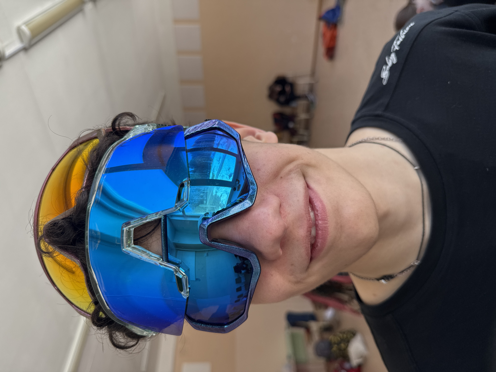 A person wearing oversized blue sunglasses and a black t-shirt is centered in the image. Part of a yellow visor is visible behind the sunglasses on the left. The person has short, curly dark hair, light skin, and a silver necklace. The background appears to be an interior space with light-colored walls and a glimpse of an open area with miscellaneous items on shelves and the floor. There is what appears to be clothing hanging near the top right. The person is facing the camera at an angle. No specific indication points to the city of Usolye, Russia.