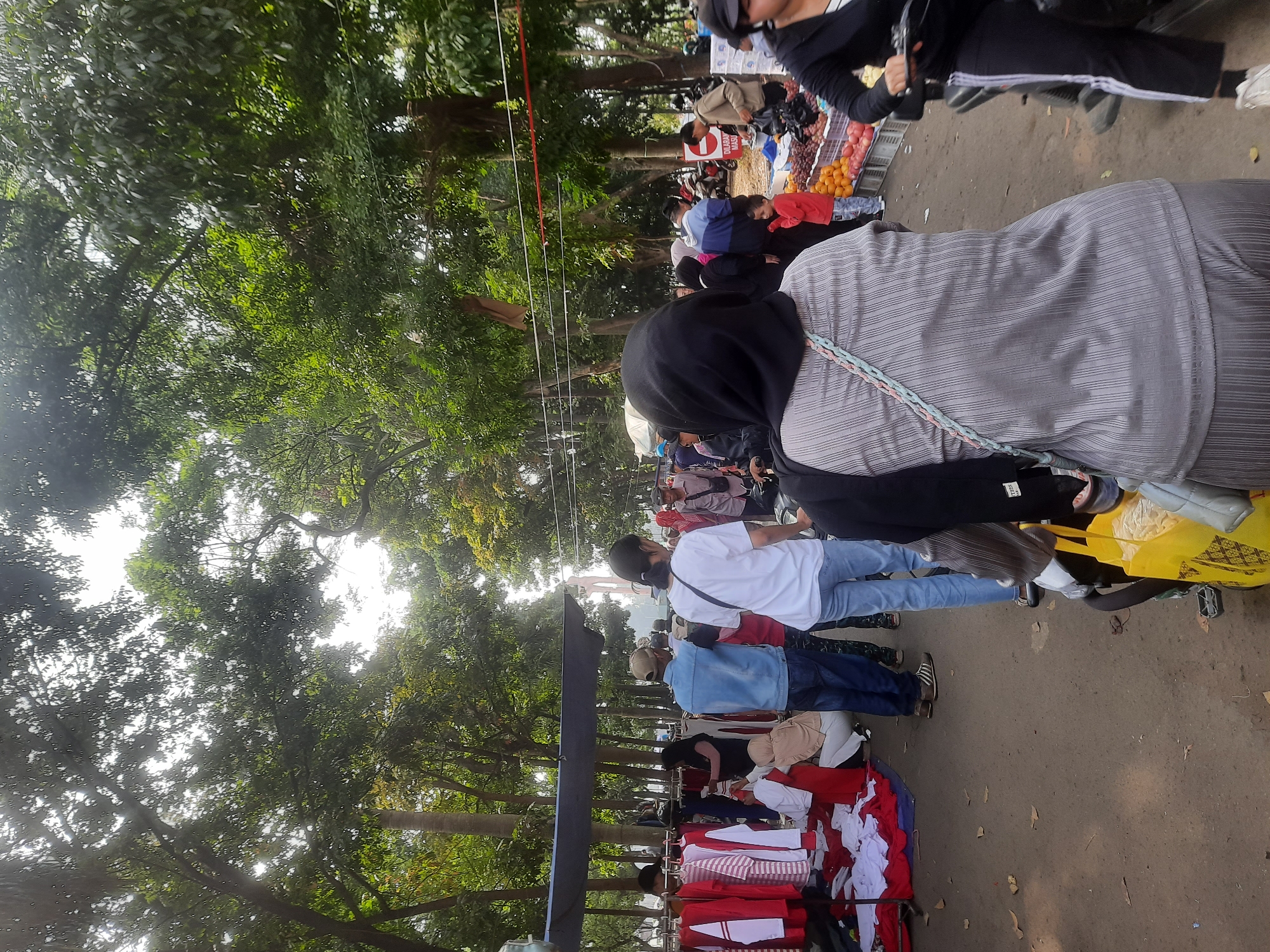 The image shows an outdoor market scene, likely in Bandung City, Indonesia, based on the style of the market and people's attire. A paved walkway is lined with market stalls. On the left, a stall displays numerous items of clothing, including red and white shirts on a rack and folded white and red fabrics on the ground. Several people are browsing or tending to the stall.
In the center of the image, a woman wearing a black hijab and a striped grey outfit is in the foreground, with her back to the camera. She appears to be riding or sitting on a motorcycle, as a yellow bag is visible in front of her. To her right, another person, also on a motorcycle, is partially visible.
Further down the walkway, more stalls are present. One stall has a variety of fruits for sale, including grapes and apples, displayed in white crates. Several people are interacting around these stalls, some appearing to be customers and vendors. The background is filled with lush green trees, suggesting a park-like setting or a street lined with vegetation. The sky is overcast, and the overall lighting indicates it is daytime.