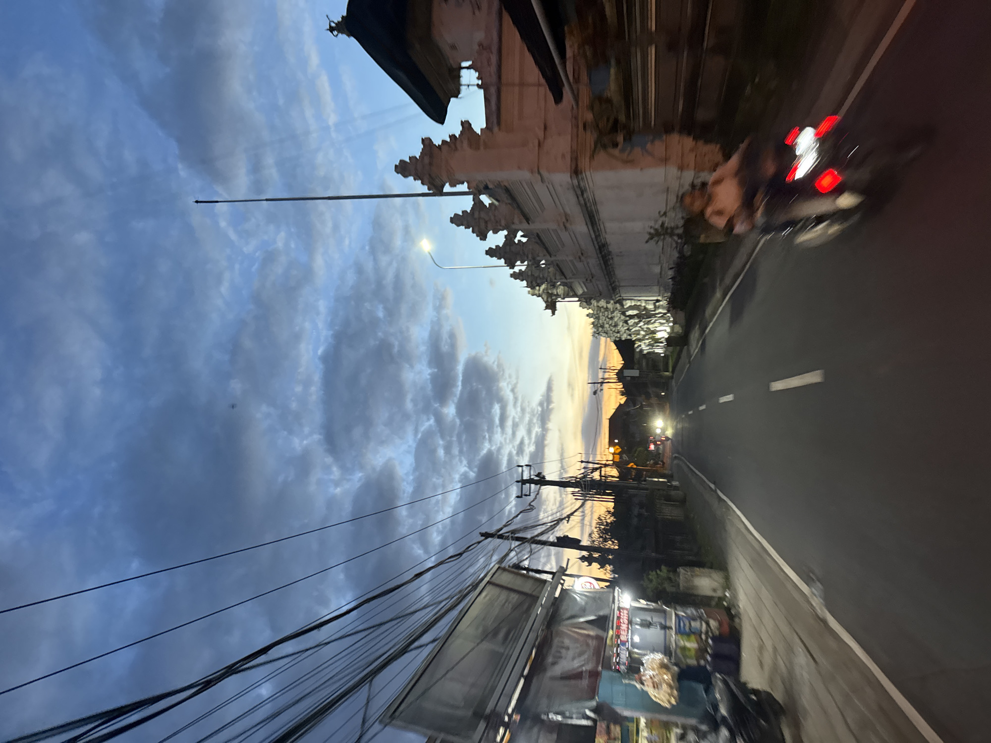 The image depicts a street scene in Kuta Selatan, Indonesia, at dusk. The sky is filled with blue and grey clouds, with a hint of orange visible near the horizon. A road runs through the middle of the frame, with a white dividing line. A blurred motorcycle with two occupants and red taillights is on the road. On the right side of the road is a traditional Balinese building with ornate carvings, which is reflected in the building. The left side of the road features a food stall with goods on display and overhead power lines. A street light illuminates the scene. The overall impression is a view from a moving vehicle or a standing person, capturing the urban landscape and atmosphere of the area.