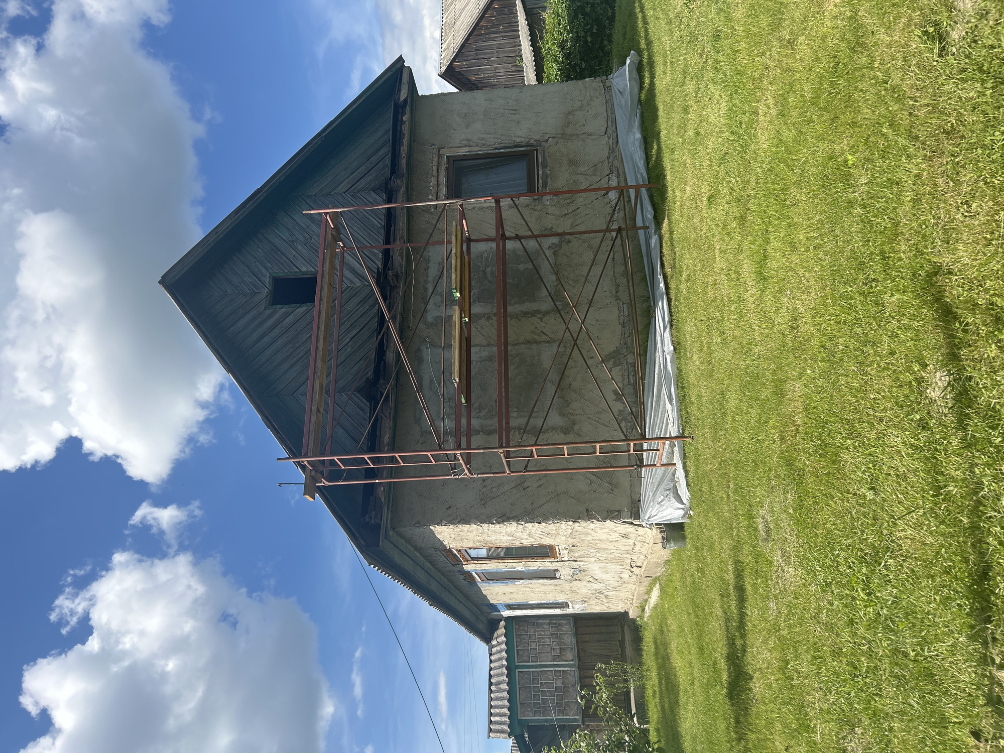 This media file depicts the exterior of a weathered building, likely undergoing renovation, situated in Vyshevychi, Ukraine. The building has a distinctively angled, dark wooden roof and walls constructed from rough, unpainted concrete. Scaffolding made of red metal pipes is erected against one side of the structure, indicating ongoing construction or repair work. A large white tarp is draped along the base of the building, further suggesting renovation activities.
The scene is set against a bright blue sky dotted with fluffy white clouds, suggesting a clear, possibly warm, day. The foreground is dominated by a lush green lawn, neatly mowed and extending towards the building. In the background, a glimpse of another wooden structure and some greenery can be seen. The overall impression is one of rural life and ongoing maintenance of older buildings. There are no people visible in the frame. The image is shot from a low angle, emphasizing the scale of the building and the sky.