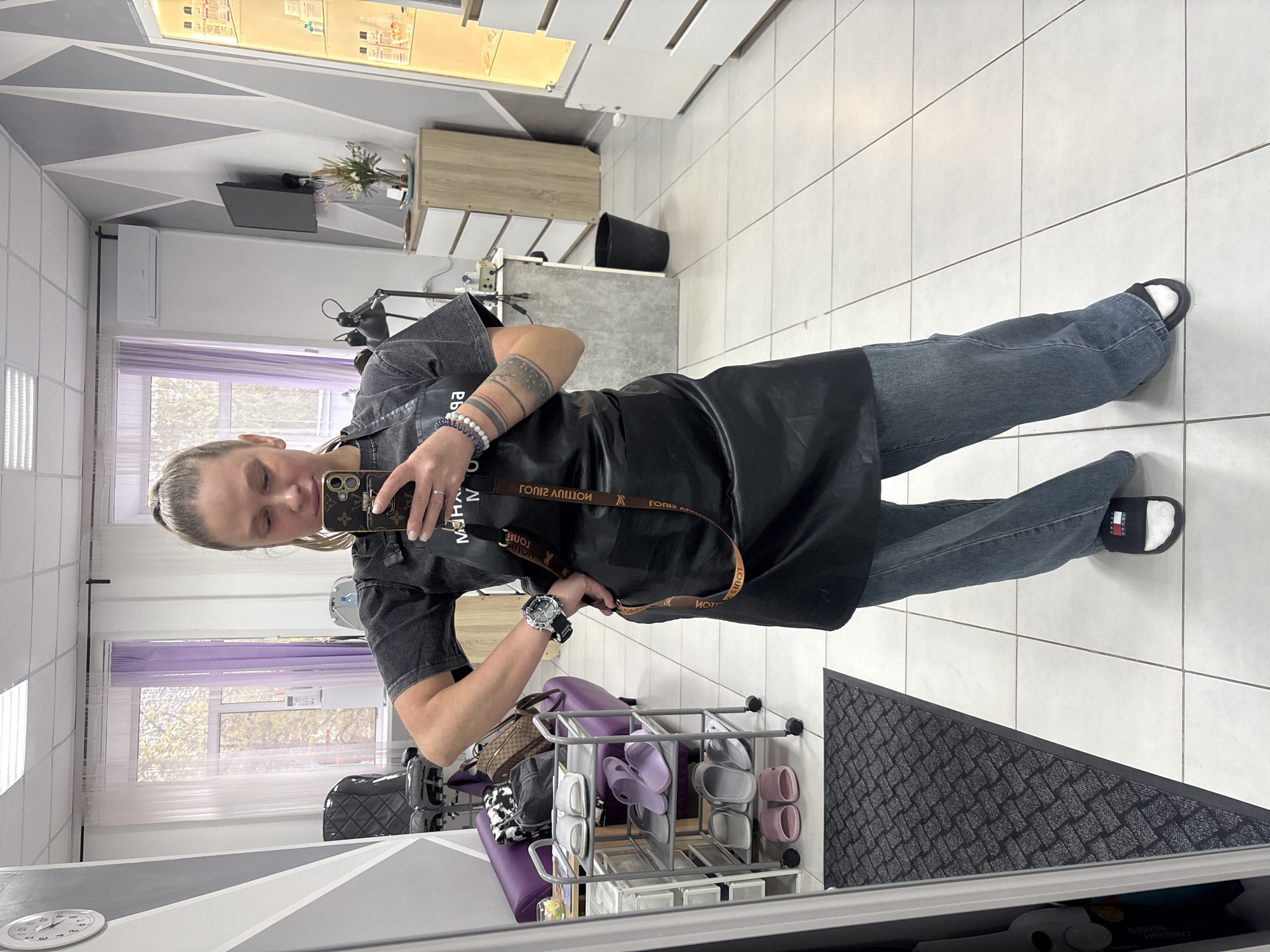 A person stands in a tiled room, taking a selfie with their smartphone. They are wearing jeans, a dark t-shirt, a black apron, and black and white slides. Their hair is up, and they have tattoos on their left forearm. A rolling cart with slippers is to their left, along with other salon furniture. The reflection shows the person is likely in a hair salon. The reflection includes a window with sheer curtains and blinds. The location is Perm, Russia.
