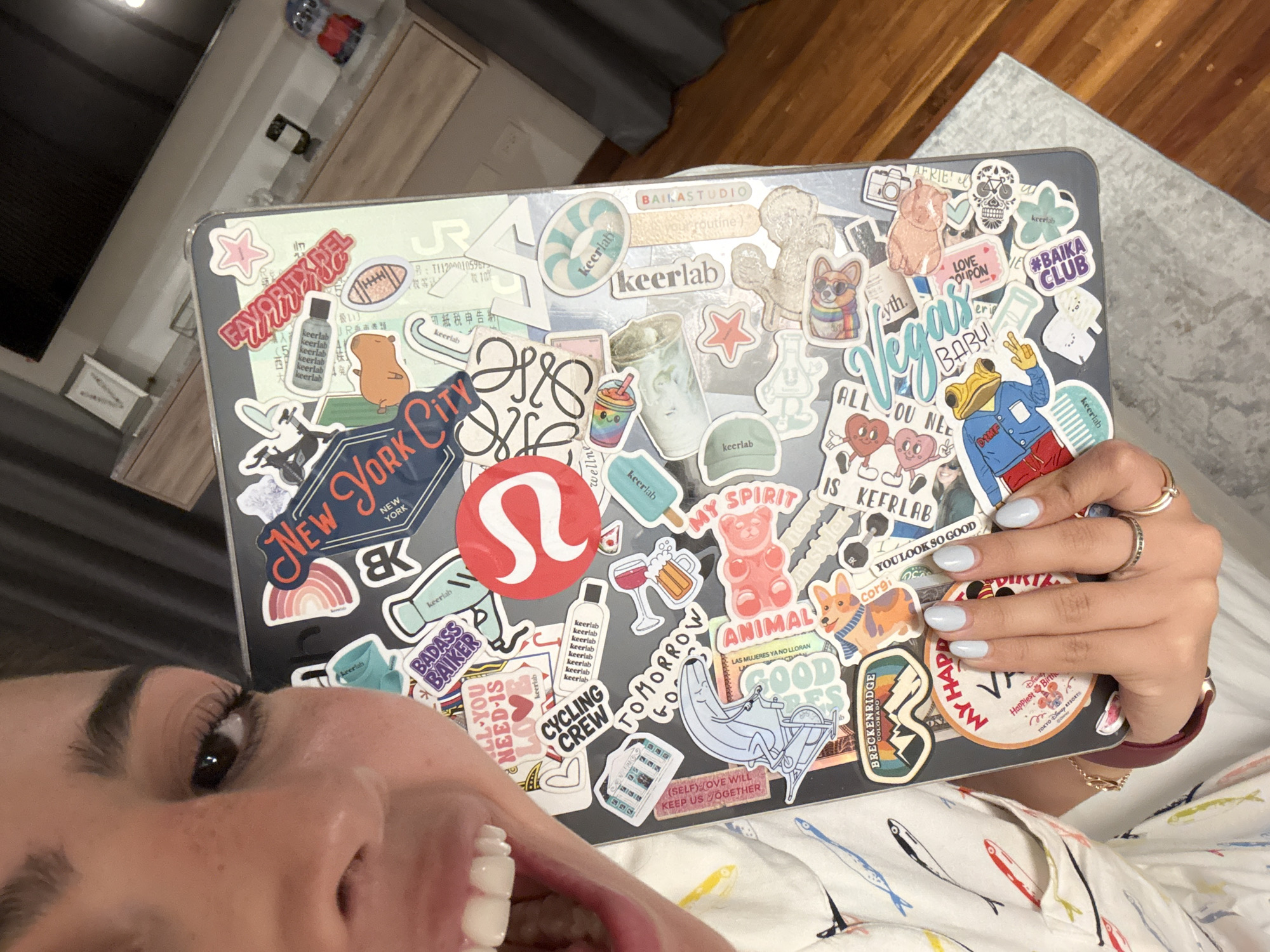 A selfie-style image features a woman holding a laptop covered in stickers. The laptop's stickers include brands and designs such as "New York City", "BAIKA CLUB", and a red Lululemon logo. The woman's left eye, part of her nose, and mouth are visible, with her teeth showing. Her nails are painted a light blue color. The background includes a kitchen or living space with a countertop and cabinets, and possibly a bed covered in a fish-patterned sheet or blanket. The image provides no information about the location in the city of San Bernardino Tlaxcalancingo, Mexico.