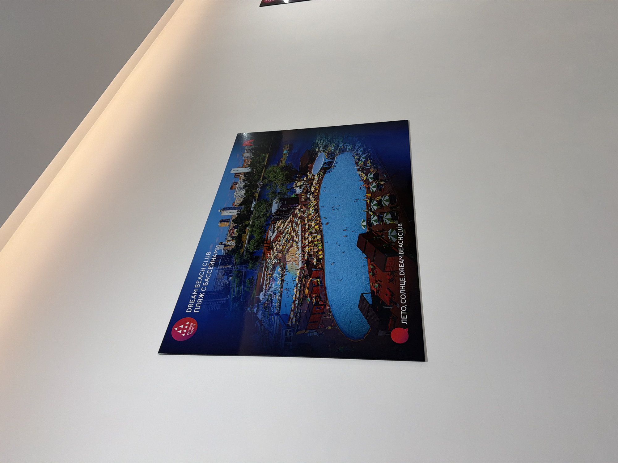 A framed poster is mounted on a white wall. The poster depicts an aerial view of a large outdoor swimming pool complex. The pool is blue and surrounded by numerous lounge chairs with umbrellas. Buildings are visible in the background, and there is a body of water, possibly a river or lake, with boats. Text on the poster is in Russian and English. The Russian text "ЛЕТО, СОЛНЦЕ, DREAM BEACH CLUB" translates to "SUMMER, SUN, DREAM BEACH CLUB". Another line of Russian text reads "ПЛЯЖ С БАССЕЙНАМИ", meaning "BEACH WITH POOLS". The logo at the top left corner has Russian text "ОСТРОВ МЕЧТЫ", which translates to "ISLAND OF DREAMS". The poster is angled, with the top left corner closer to the camera than the bottom right. The wall is illuminated from above with warm light.