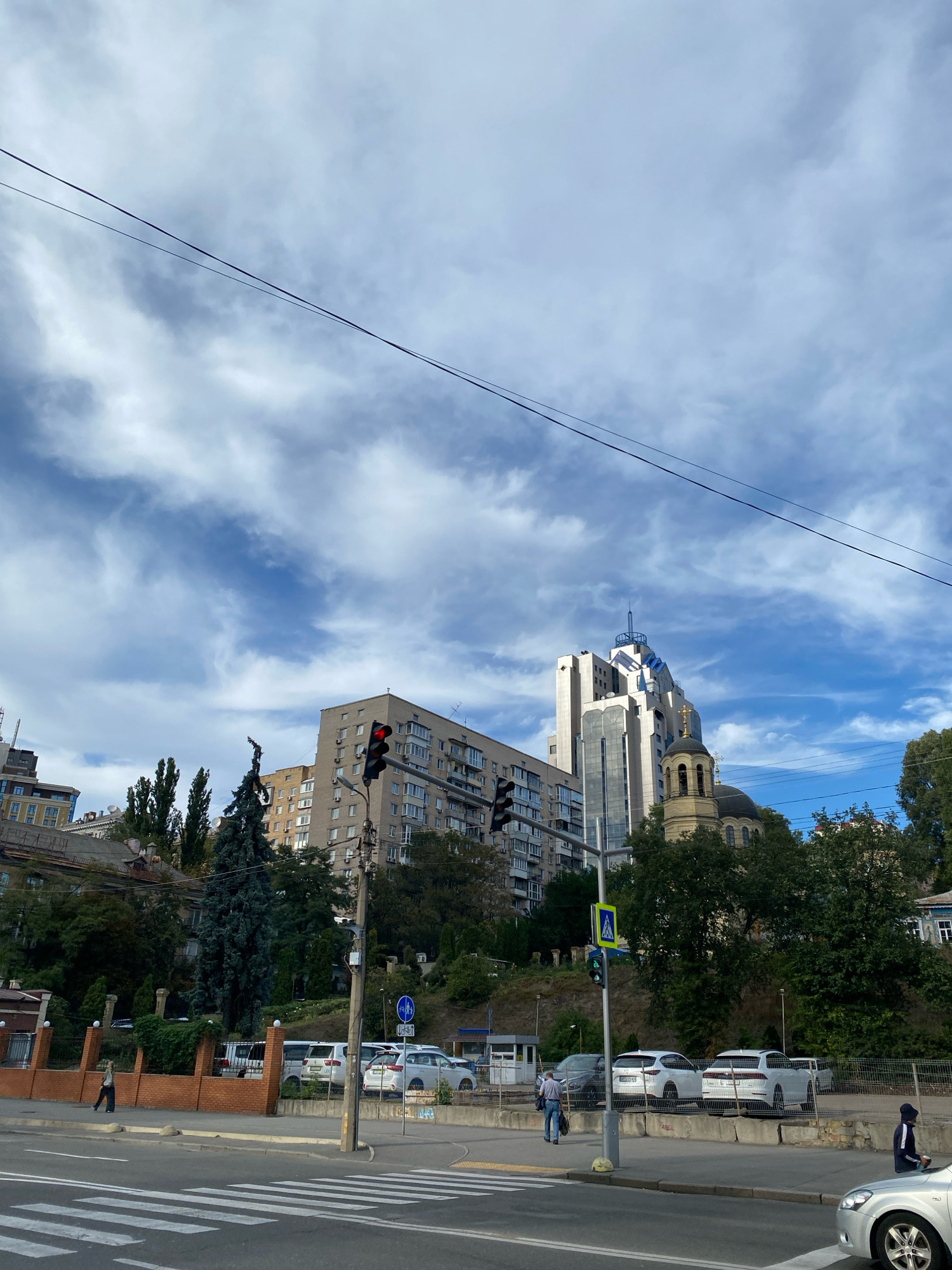 The image shows a city street scene with buildings, cars, and pedestrians under a cloudy sky. A traffic light is visible in the center-left, showing a red signal. Several cars are parked along the side of the road. A pedestrian is walking on the sidewalk. In the background, there are multi-story buildings and some trees. The overall environment suggests an urban setting.
