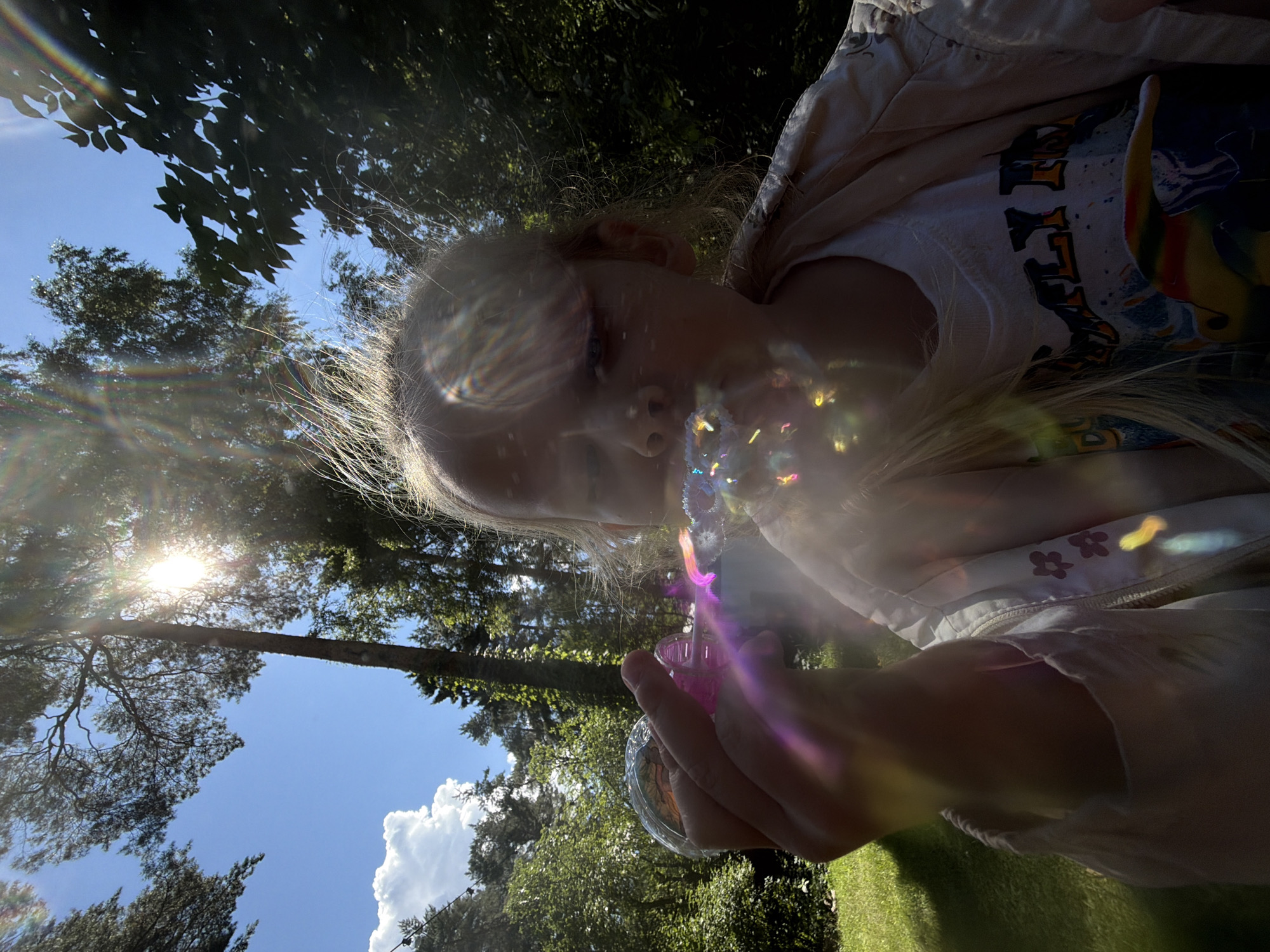 The media file shows a close-up, angled shot of a young child blowing bubbles outdoors. The child, with blonde hair, is seen in profile, looking downwards towards a bubble wand and the bubbles being formed. The bubbles are iridescent and catch the light, creating rainbow-like reflections across the image, especially on the child's face and hair. The child appears to be wearing a light-colored jacket over a t-shirt with some graphic print.
The setting is a sunny outdoor environment, likely a park or backyard, with lush green trees and grass visible. The sky is blue with a few white clouds. The sun is bright, causing lens flare and creating a radiant effect in the upper left quadrant of the image, shining through tree branches. The time of day appears to be daytime, likely late morning or afternoon, given the bright sunlight. The overall mood seems playful and joyful, typical of a child engaged in an activity like blowing bubbles on a pleasant day.
The orientation of the image is rotated, with the child's head pointing downwards relative to the typical upright orientation. The location context provided is Udelnaya, Russia. There are no clearly legible words or text visible in the image.