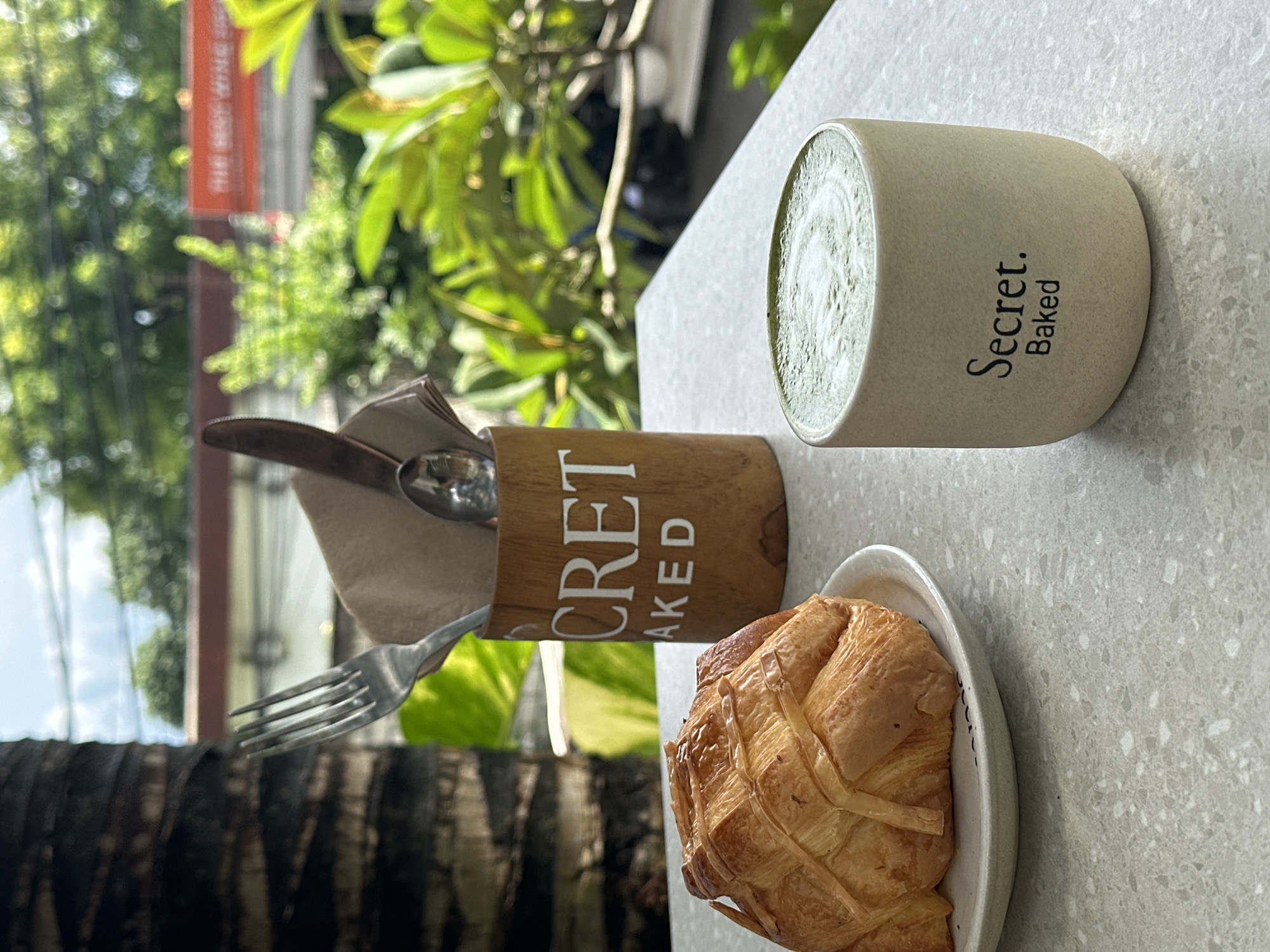 Here is a description of the image:
The image captures a breakfast scene at Secret Baked, located in Nusa Penida, Indonesia. The setting is an outdoor table with a view of lush greenery. A matcha latte in a light beige mug with "Secret. Baked" printed on it sits on the table. Beside the mug, there's a wooden container holding a fork, spoon, and napkins. In front of these items, a croissant is served on a small white plate with dark accents. The composition of the shot and the angle of the food suggest it's likely a photo taken to document a meal at this specific location.