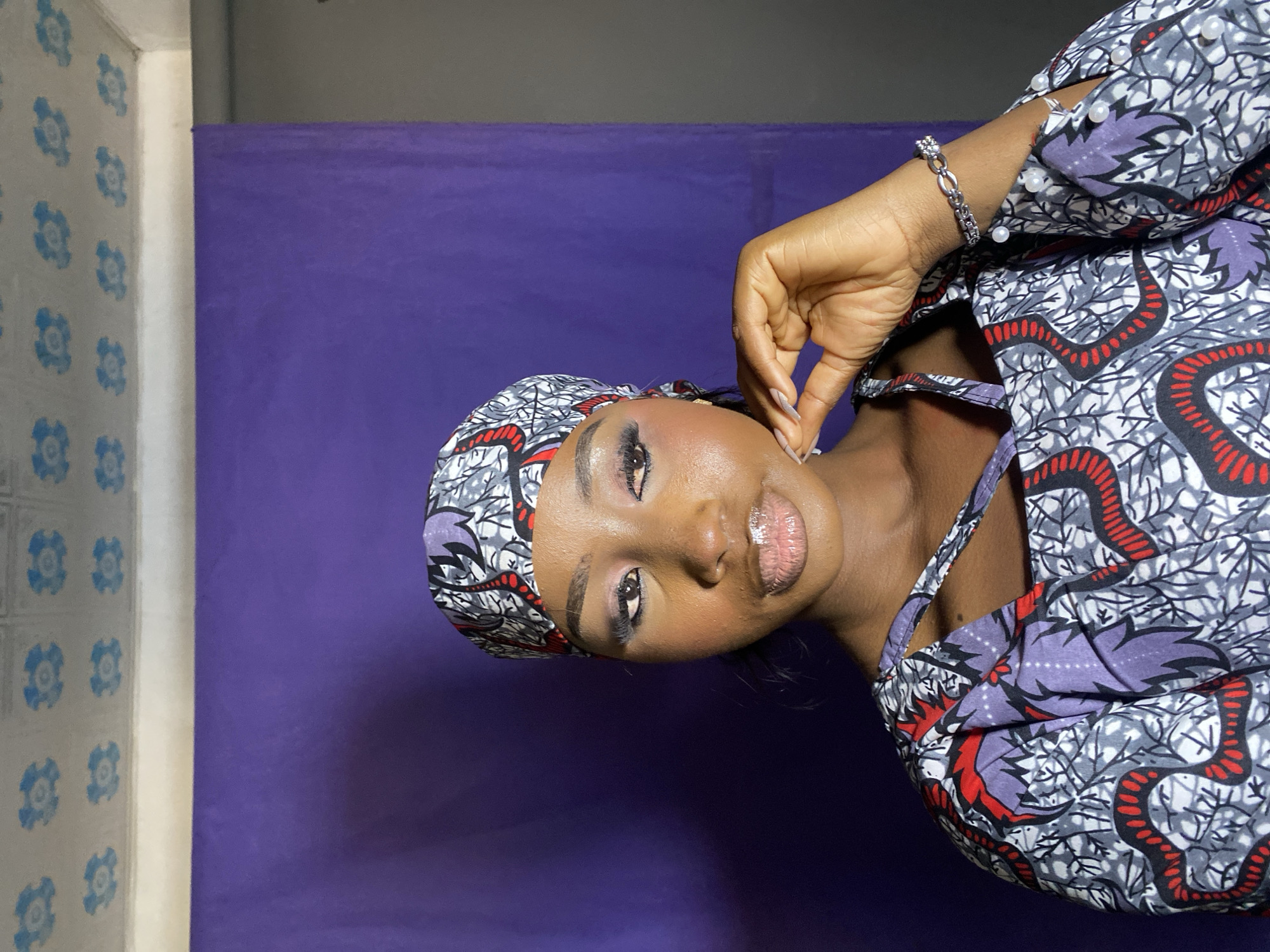 A woman with brown skin, wearing a patterned headscarf and dress, poses in front of a backdrop of blue and white wallpaper to the left and a solid purple screen behind. Her left hand is raised to her face. She also is wearing a silver bracelet. The location is Gura Top, Nigeria.