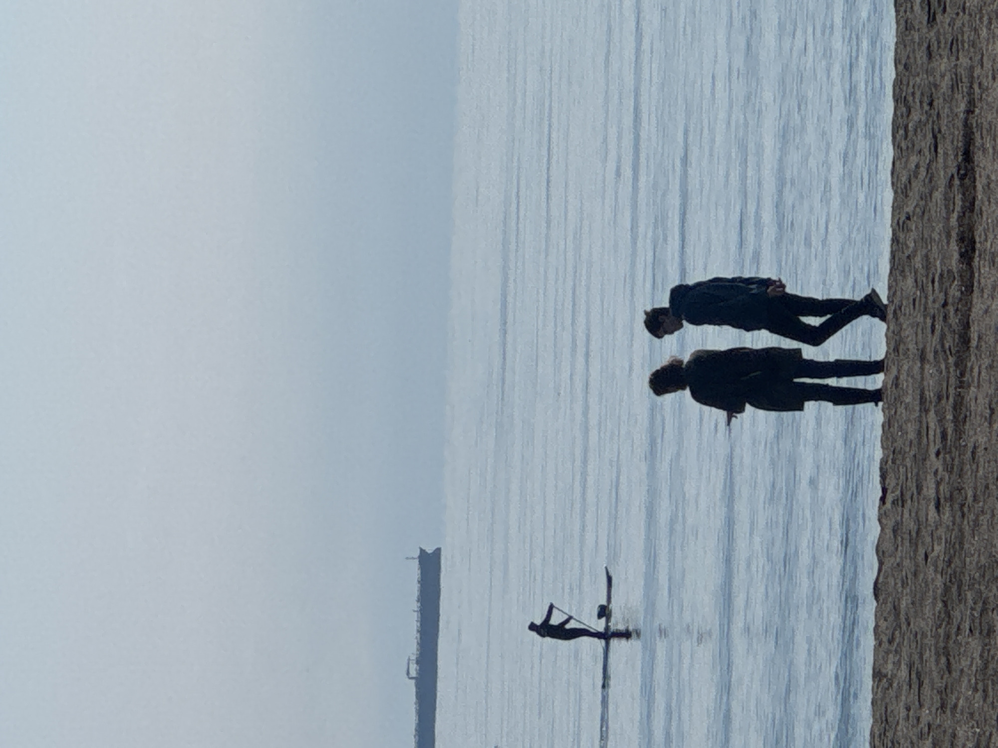 The image shows a scene at Burgas beach, Bulgaria. Two people are standing on a concrete pier or breakwater on the right side of the frame. They are both wearing dark clothing and appear to be looking out at the water. In the water, there is a windsurfer, and in the distance, a pier or a large structure is visible in the hazy background. The water has a choppy texture, and the sky is overcast.