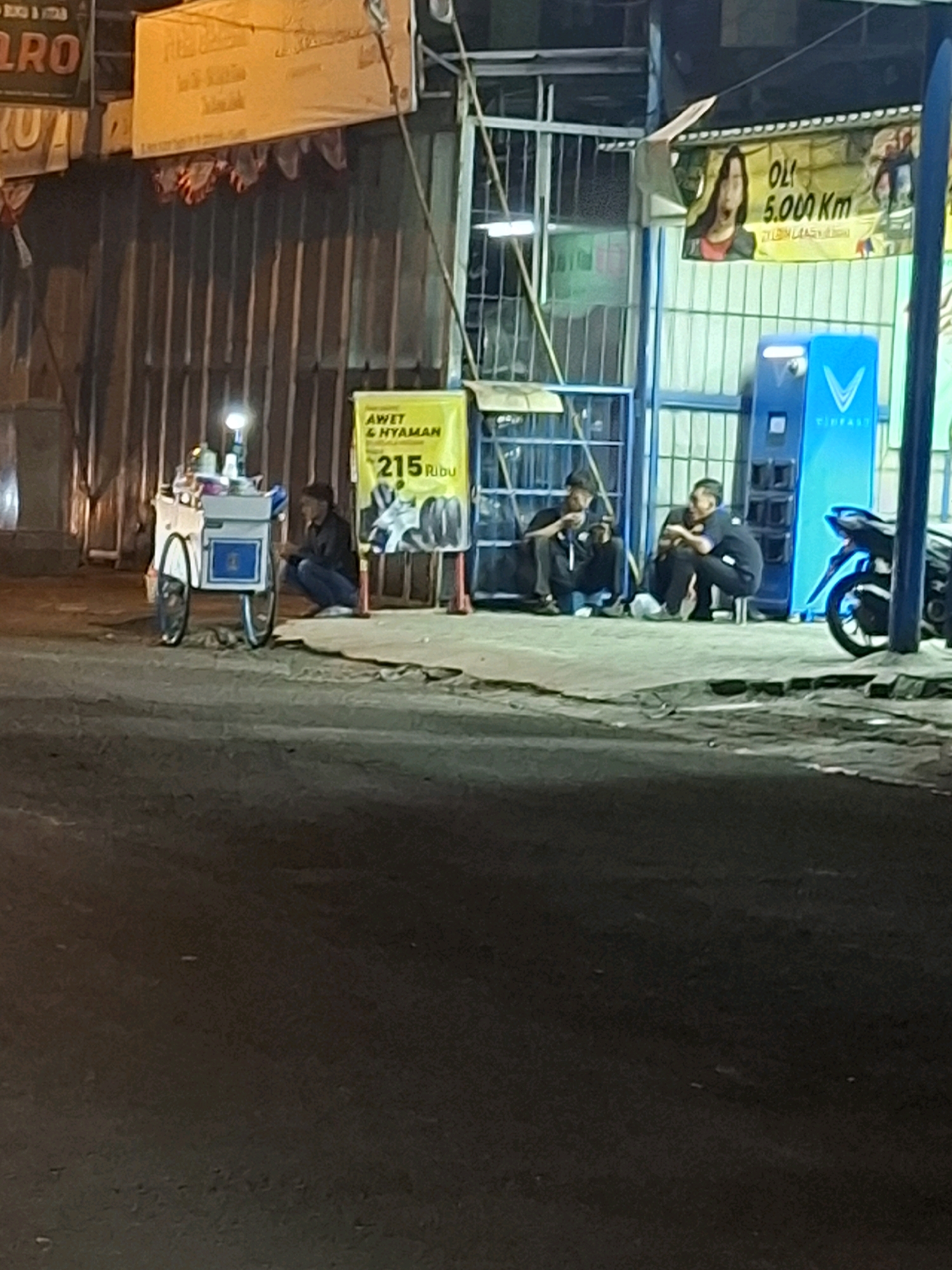 The image captures a street scene in Cianjur, Indonesia, at night. A man is crouched beside a mobile ice cream cart, which is equipped with a light and various containers. In the background, several other individuals are seated near a building with a metal corrugated facade and blue-tinted windows. One person is holding a smartphone. A blue electric charging station is visible to the right of the individuals. A motorcycle is parked to the far right. Several banners hang above the building's entrance, one of which displays text in Indonesian and an image of a woman, possibly an advertisement. The overall lighting suggests it is after dusk, with artificial lights illuminating the area.