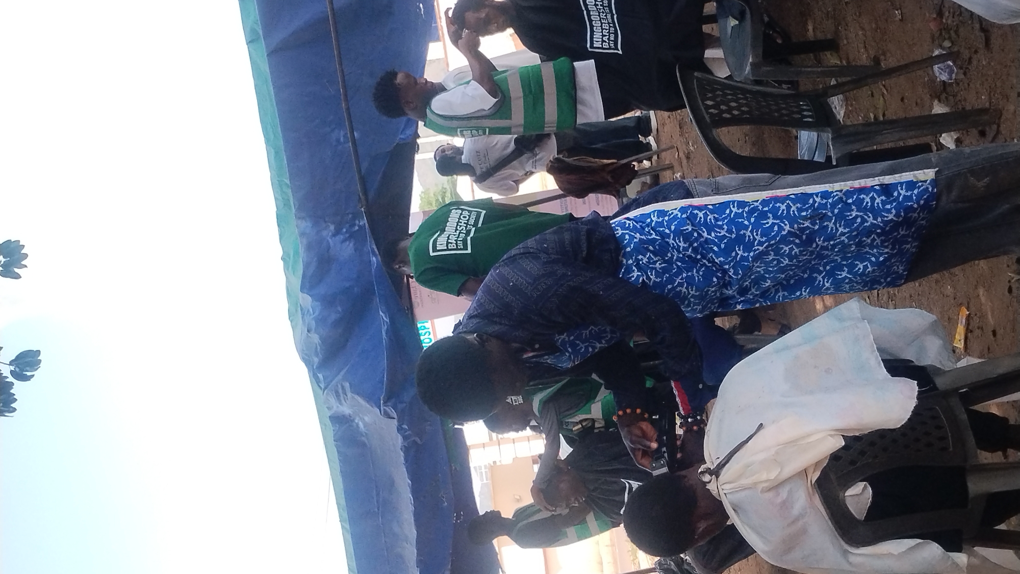 The image shows a group of people gathered under a blue and white canopy tent, possibly outdoors. Some are sitting in plastic chairs. Several men are wearing shirts that identify a barbershop: "KINGDONS BARBERSHOP, SAY NO TO A 1981 SS SOCIETY". One person wears a green and white safety vest. The location is possibly in Jos, Nigeria, based on the OCR text identifying the barbershop. A person in a white cape appears to be getting a haircut. The ground appears to be dirt.