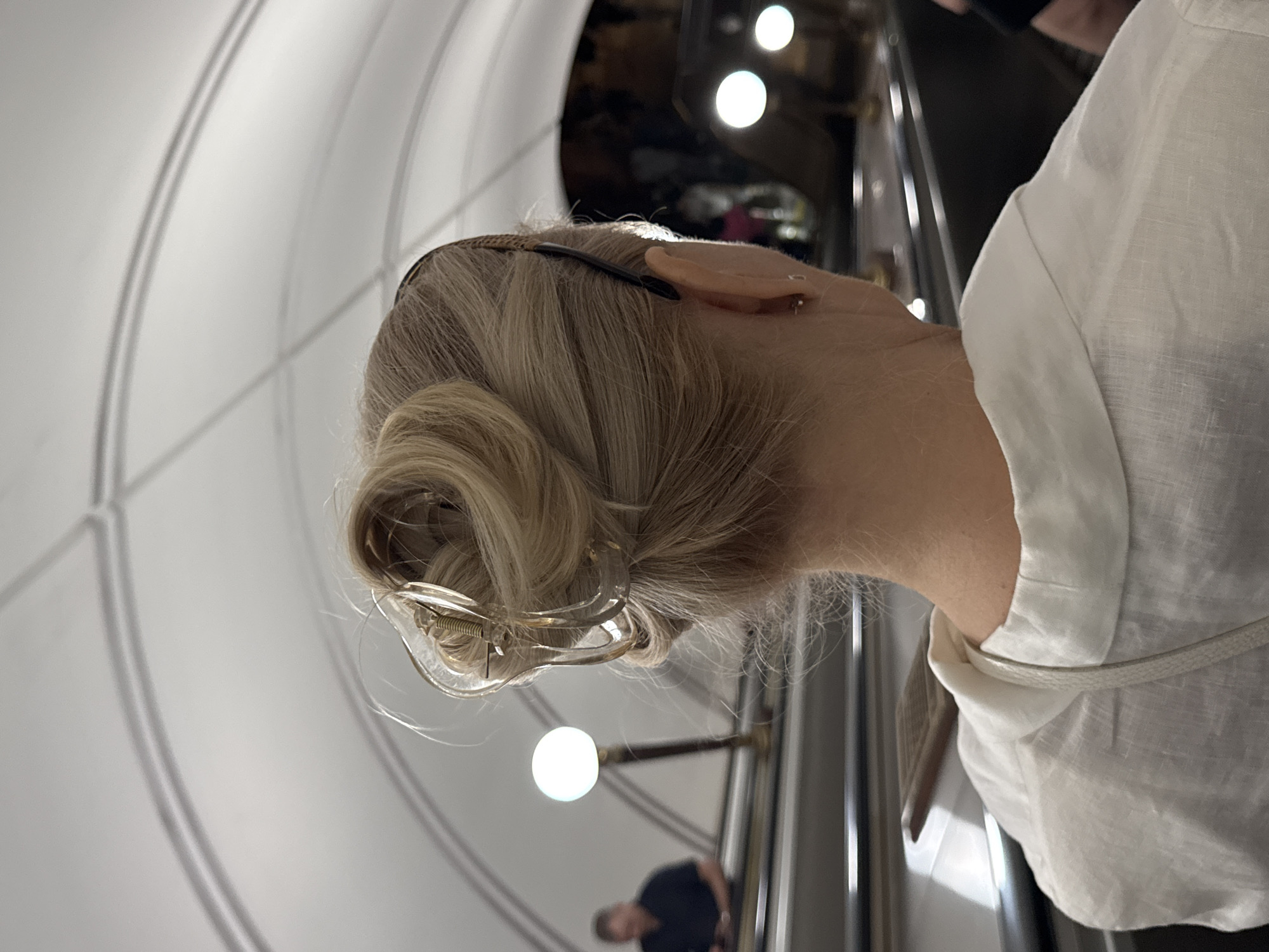 The image shows the back of a blonde woman's head, wearing a white linen shirt and a large, clear plastic hair clip. She has a small silver earring in her left ear. The background appears to be an indoor public space, possibly a metro station, with curved, white ceilings and bright lights. On the left, an escalator is visible with a man in a dark shirt descending. To the right, another escalator is seen with blurred figures and lights. The overall ambiance suggests a bustling urban environment. While not explicitly identifiable, the architectural style and lighting are consistent with Moscow's metro stations.