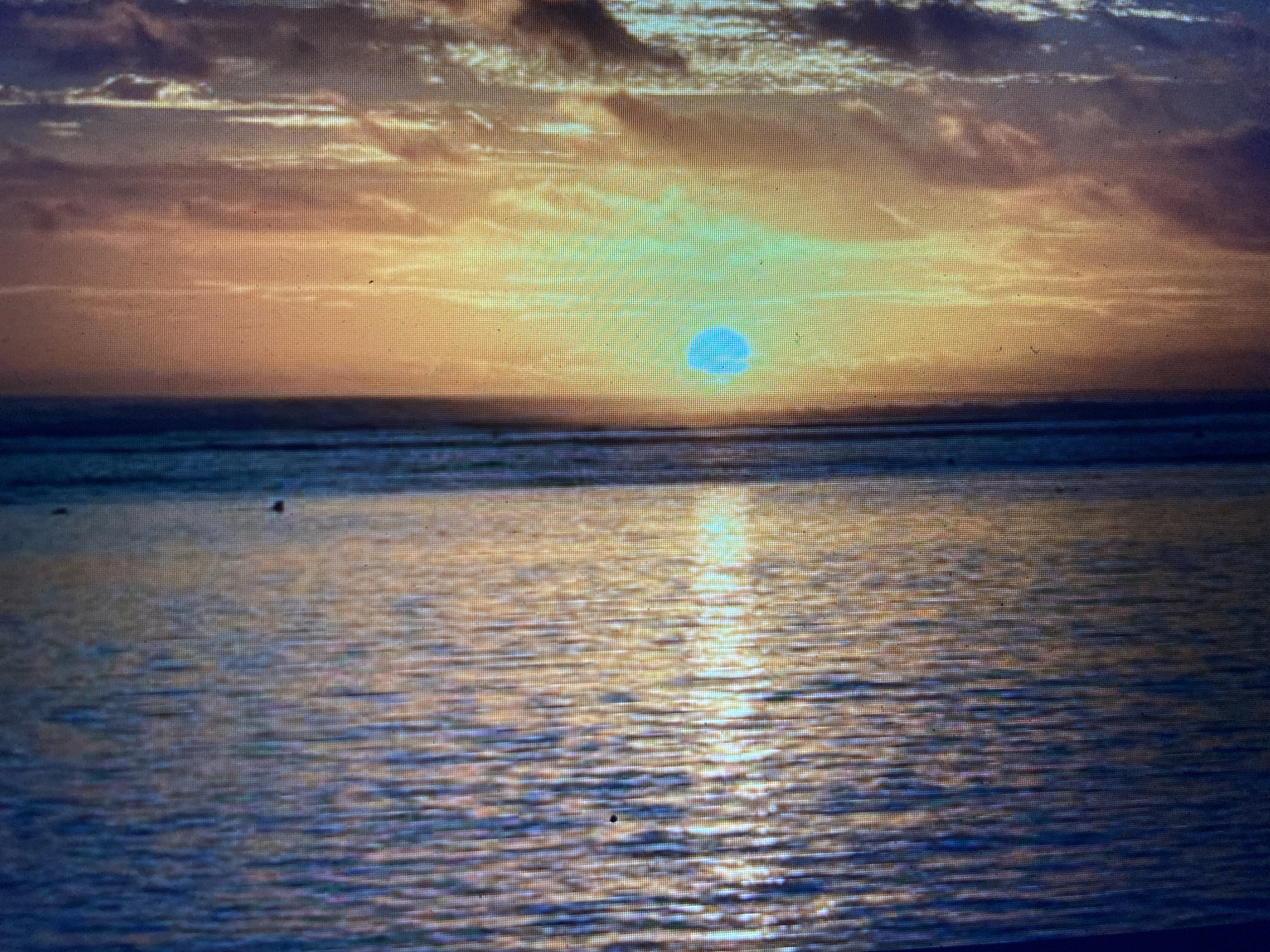 A digitally rendered image depicts a sunset over a body of water, with the sun appearing as a bright cyan orb. The sky is a gradient of orange and yellow, with streaks of darker clouds. The water is choppy and reflects the colors of the sky and the sun. The image is pixelated, suggesting it is a screenshot or a low-resolution digital display. There are no people or specific landmarks visible, making it impossible to determine a specific location within Girijaya, Indonesia.