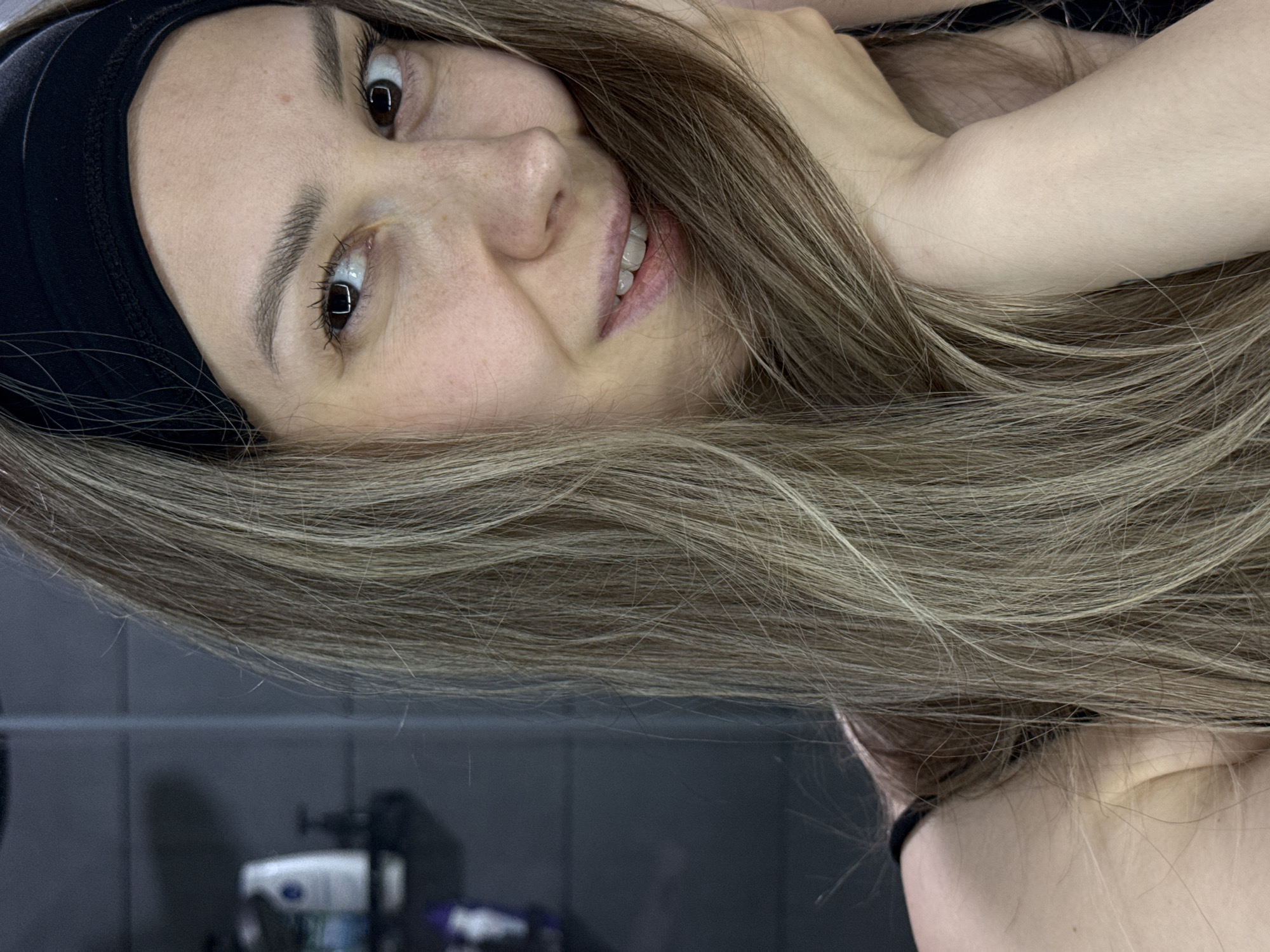 A light-skinned woman with long, light-brown and blond-streaked hair is smiling at the camera. She is wearing a black headband. The background is a dark, tiled wall with blurred objects on a shelf. There is no explicit information to confirm this image was taken in Chaikovsky, Russia.