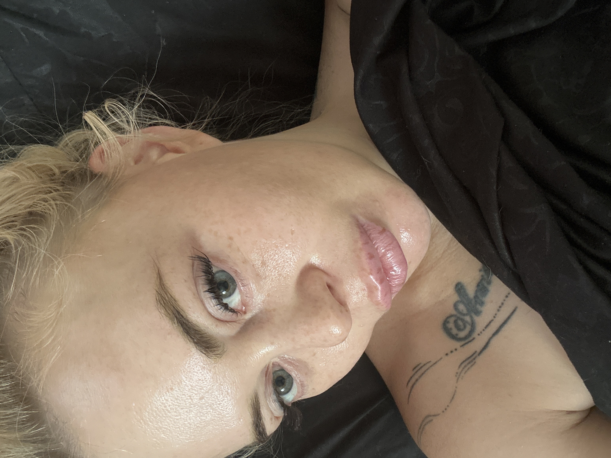 A close-up, high-angle selfie shows a blonde woman with fair skin and freckles, lying down and looking towards the camera. She has light blue eyes, dark eyebrows, and long, dark eyelashes. Her lips are full and appear to be glossy. Her hair is pulled up in a messy bun, with some strands loose around her face and ears. She is wearing a black garment that covers her chest and shoulder, on which a tattoo is visible. The background is a dark, solid color, possibly a black sheet or blanket. The lighting is soft, creating a slight sheen on her skin. The image appears to be taken indoors.