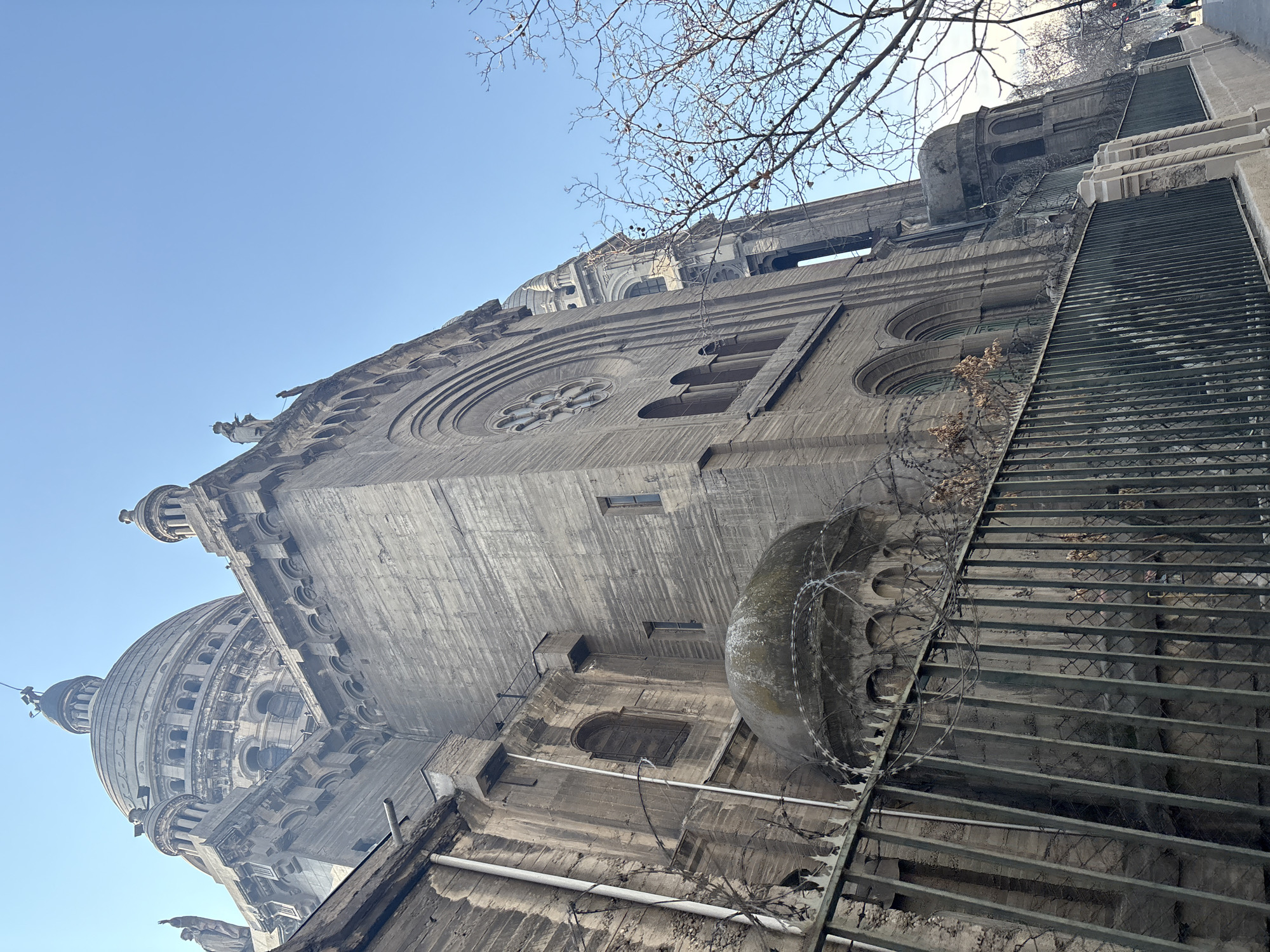 The image shows a low-angle view of the Basilica of the Sacred Heart of Jesus in Santiago, Chile, on a clear, sunny day. The basilica's imposing stone facade dominates the frame, with its grand dome and detailed architectural features. A wrought-iron fence topped with barbed wire runs along the foreground, separating the viewer from the building. Bare tree branches are visible on the right side of the image, suggesting it might be winter. The sky is a vibrant blue.