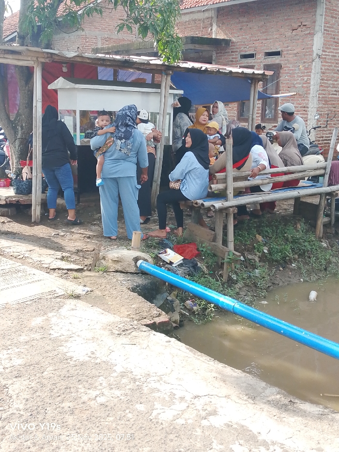 A group of women and children gather under a makeshift structure. Some are seated on a bamboo bench, while others stand nearby. Most of the women wear headscarves. A man sits on a motorcycle in the background. In the foreground, a blue pipe crosses over a small body of water. The structure appears to be near a brick building with exposed brickwork. The setting appears to be a roadside or gathering place.