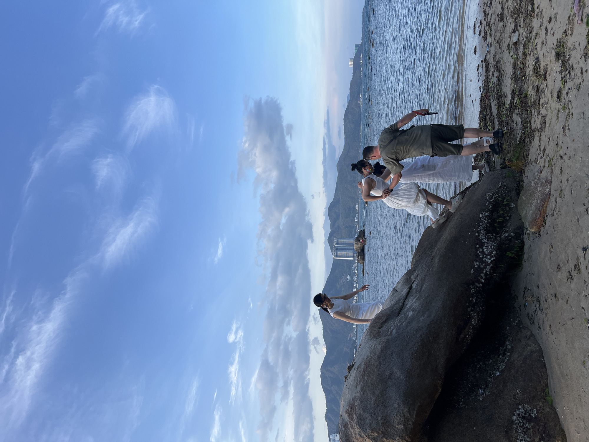 The image depicts a coastal scene in Bac Nha Trang Ward, Vietnam, featuring four individuals on a rocky and sandy shoreline under a bright, partly cloudy sky. The photograph appears to be taken during the late afternoon or early evening, given the clear blue sky with scattered white cirrus clouds and some larger, grey clouds near the mountainous horizon.
In the left foreground, a man wearing a white top and light-colored pants is poised on a large, dark rock, extending his arms as if balancing or performing. Further to the right, closer to the water, a man in a dark green shirt and shorts is seen holding a smartphone, seemingly capturing a moment. He stands beside two women, both dressed in white attire. One woman, with her hair pulled back, is positioned near the man with the phone, appearing to hold his hand or arm. The other woman in white stands slightly further right, closer to the water's edge.
The immediate setting includes various large rocks, some covered with barnacles, leading to a stretch of sand mixed with dark seaweed along the water. The water itself shows gentle ripples, extending towards a distant coastline dotted with high-rise buildings and receding mountains. The overall activity appears to be a coordinated pose or a photoshoot in a picturesque natural environment. There is no visible text in the image.