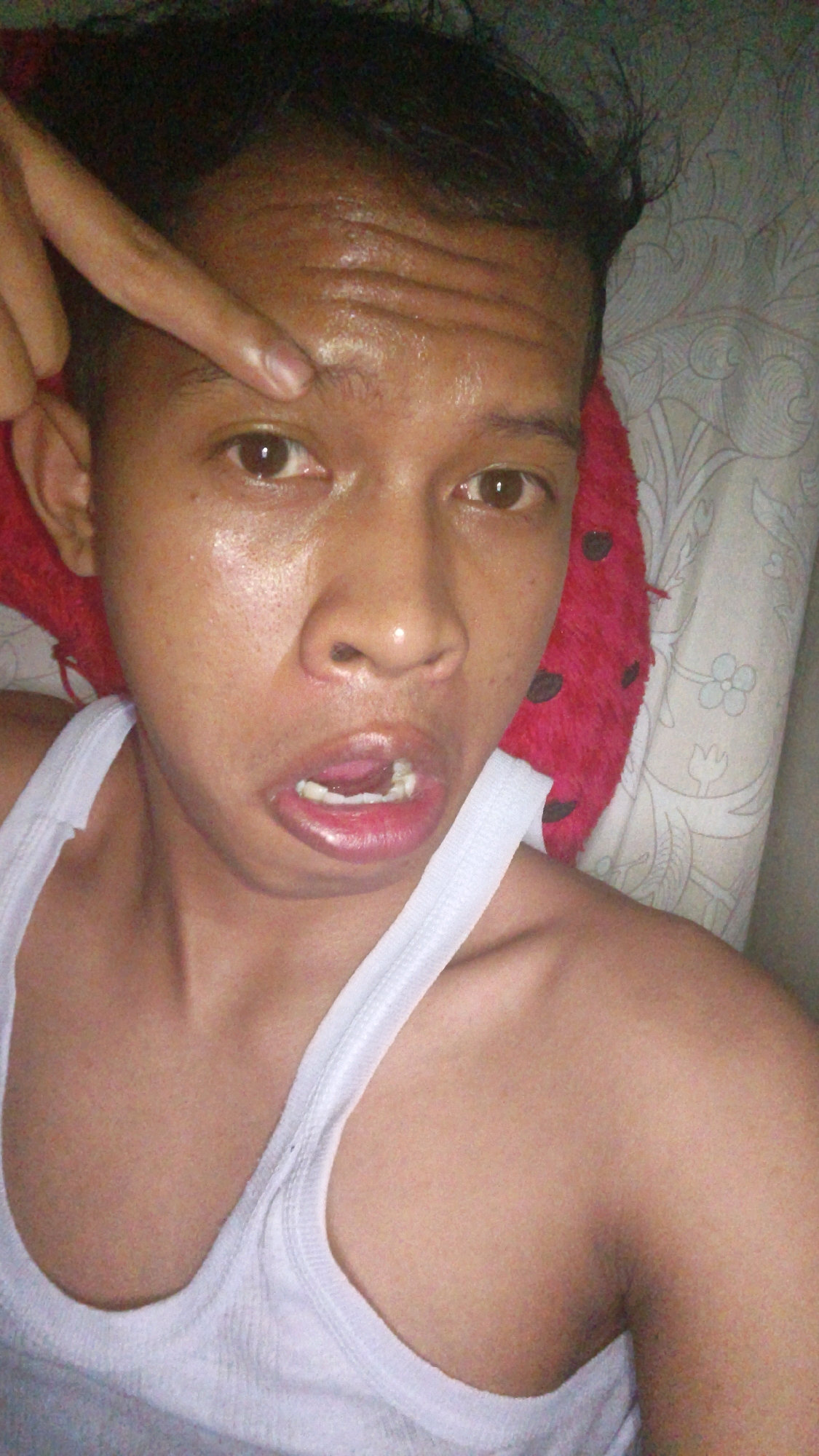 A close-up selfie shows a person from the chest up, facing the camera. The person has dark hair and brown eyes, and is wearing a white tank top. Their mouth is slightly open, revealing teeth, and their forehead appears shiny. A finger is pointing at the person's forehead. The background is out of focus, with what appears to be a pillow with a red, fuzzy texture and a patterned textile. The image is poorly lit and has a grainy quality. No specific location in Pundungsari, Indonesia is discernible from the image.