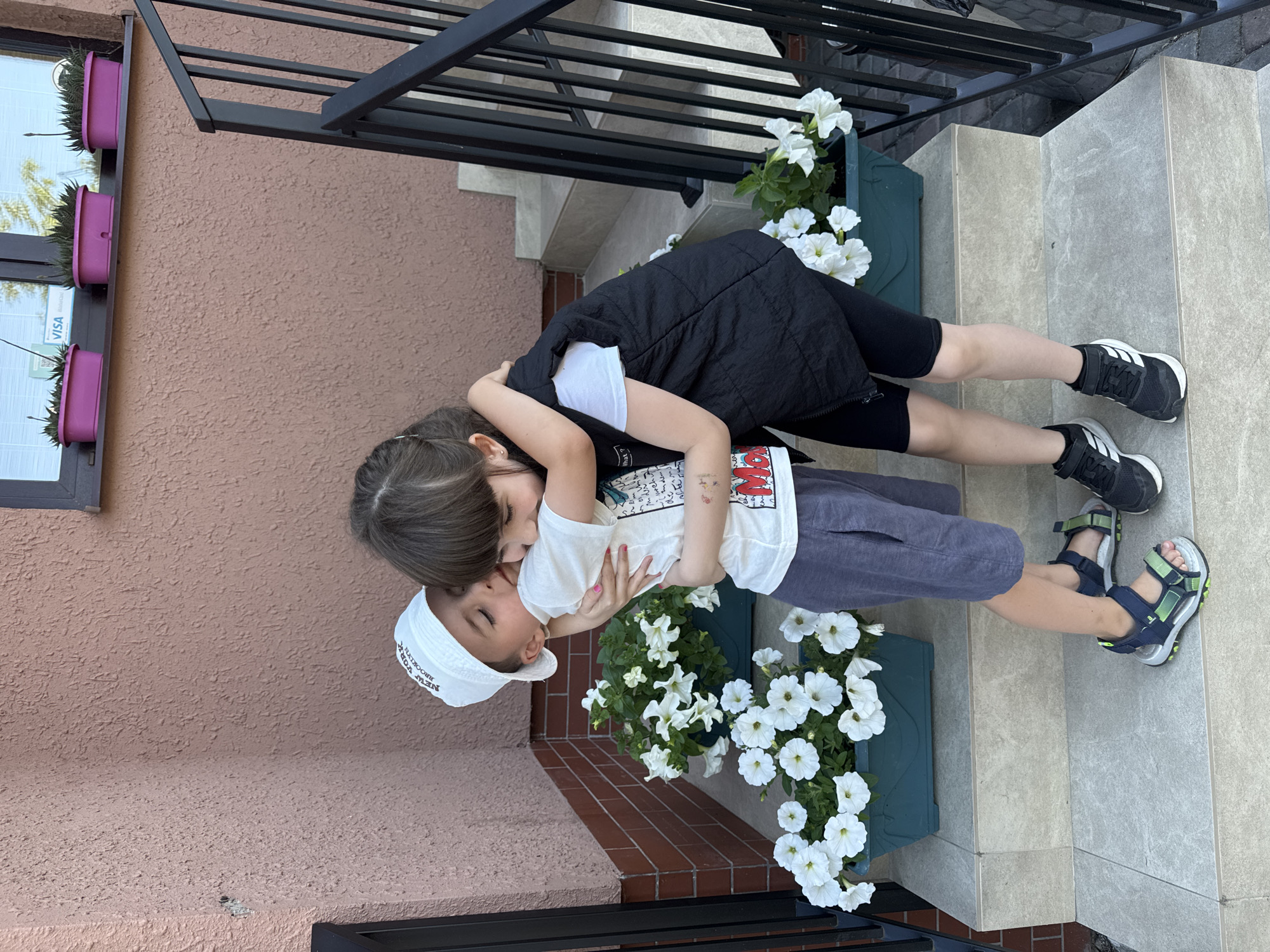 Two children, a boy wearing a white cap and a white t-shirt with a graphic print and blue shorts, and a girl wearing a white t-shirt and a black vest with black shorts, are hugging on a set of concrete stairs in front of a textured pink wall. The boy is kissing the girl on the cheek. White petunias are planted in green rectangular planters on the stairs. A black metal railing is visible to the right. The background shows a window with three purple planters containing plants. The image appears to be taken outdoors on a sunny day. No specific identifiable landmarks or indications place this scene in Kolomyia, Ukraine.