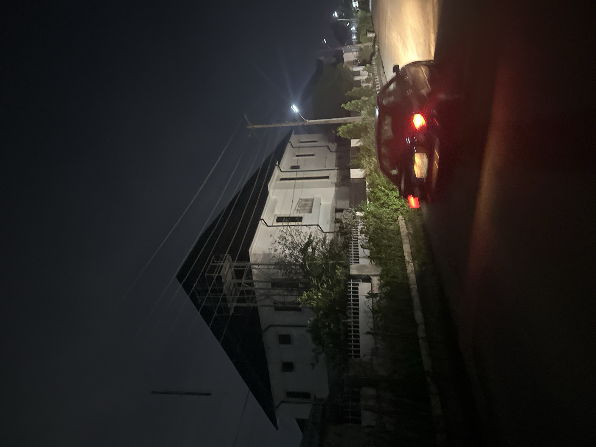 The image depicts a nighttime scene in a residential area of Abuja, Nigeria. A white, multi-story building with a dark, sloped roof dominates the left side of the frame. The building features several windows and a balcony. In front of the building, there is a white metal fence and dense vegetation. A black sedan is driving away from the camera on a paved road, with its red taillights illuminated. Streetlights are visible along the road, casting a warm glow. The sky is dark, suggesting late evening or night. The overall environment appears to be a quiet, well-maintained suburban street.