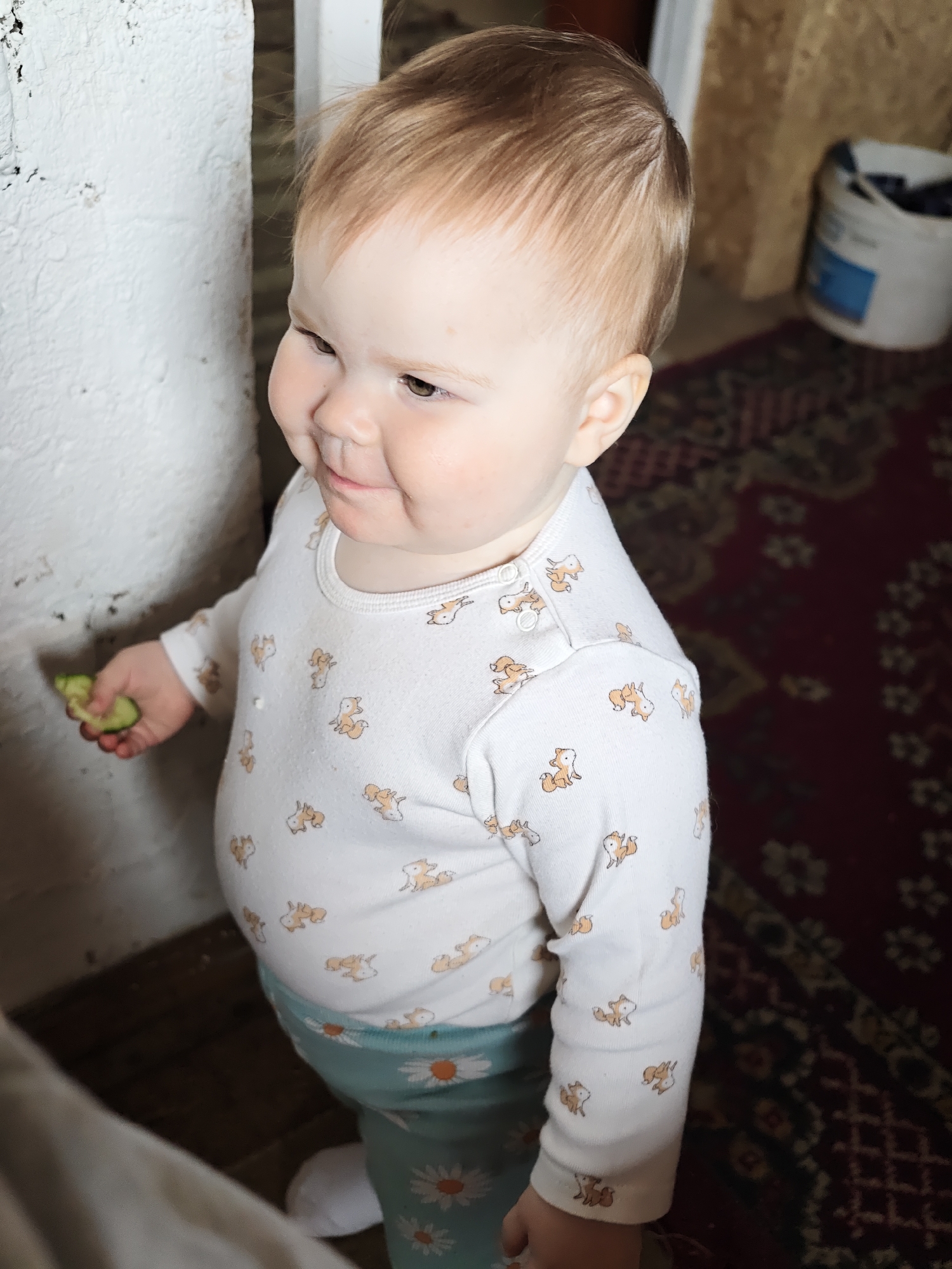 A toddler stands indoors, holding a green piece of food in their left hand. They wear a white long-sleeved shirt patterned with fox-like figures and blue pants with a white daisy print. The toddler is facing towards the left of the frame with their head turned slightly toward the same direction. To the toddler's left is a textured white wall. The background is blurred, but a red patterned rug is visible on the floor, and a white bucket is on a table or shelf. The location is Malyi Bukor, Russia.