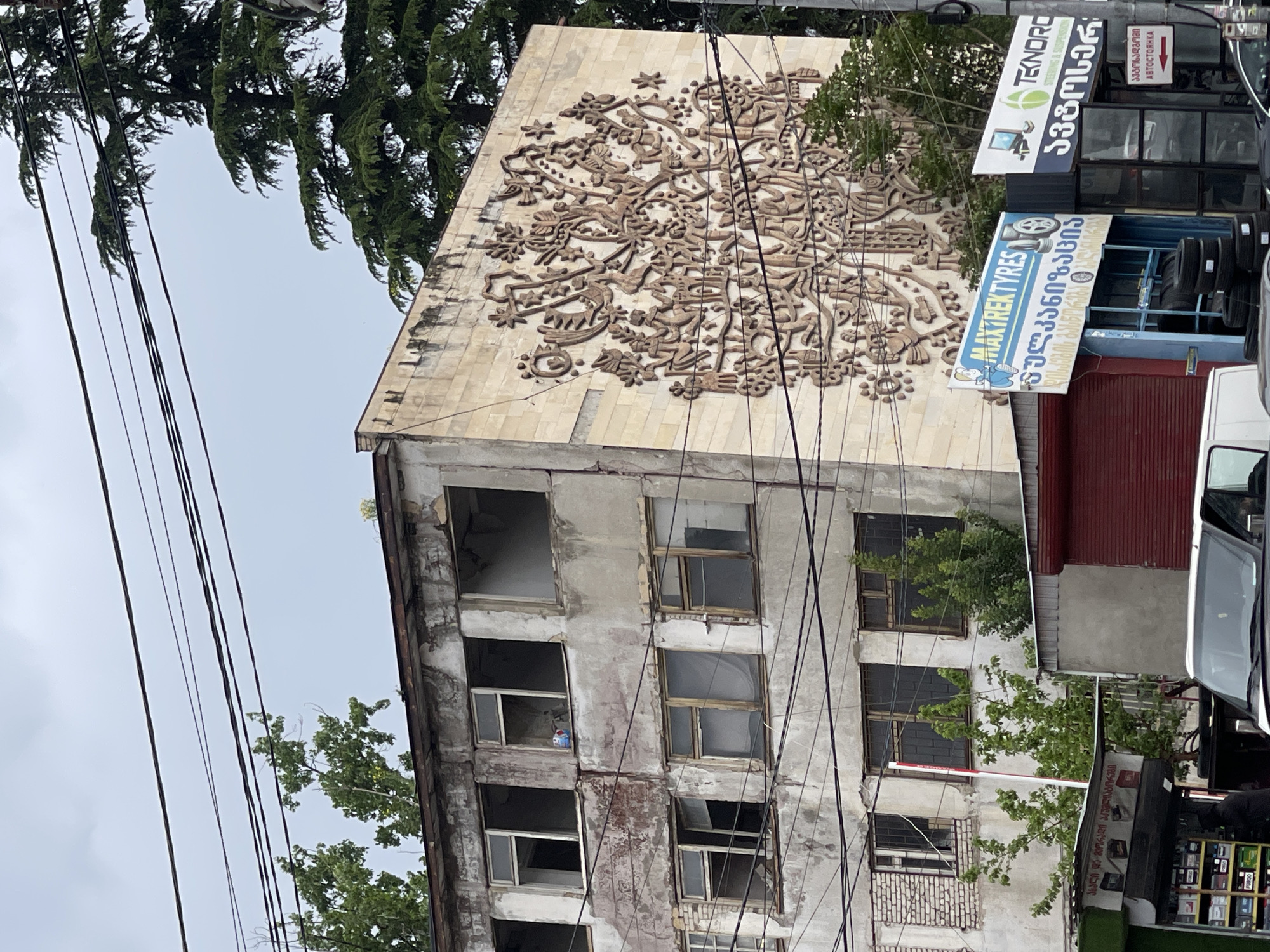The image shows a rundown building in Tbilisi, Georgia, adorned with a large, circular bas-relief on its upper facade. The bas-relief features a complex, stylized design. The building's windows are largely empty and lack panes, suggesting abandonment or disrepair. A collection of signs for local businesses, including "TEKNOPO," "MAYREKTYRES," and another sign in Georgian script, are visible attached to a newer structure adjacent to the old building. Trees partially obscure the view of the building and are also visible along the top of the image. Power lines run across the foreground.