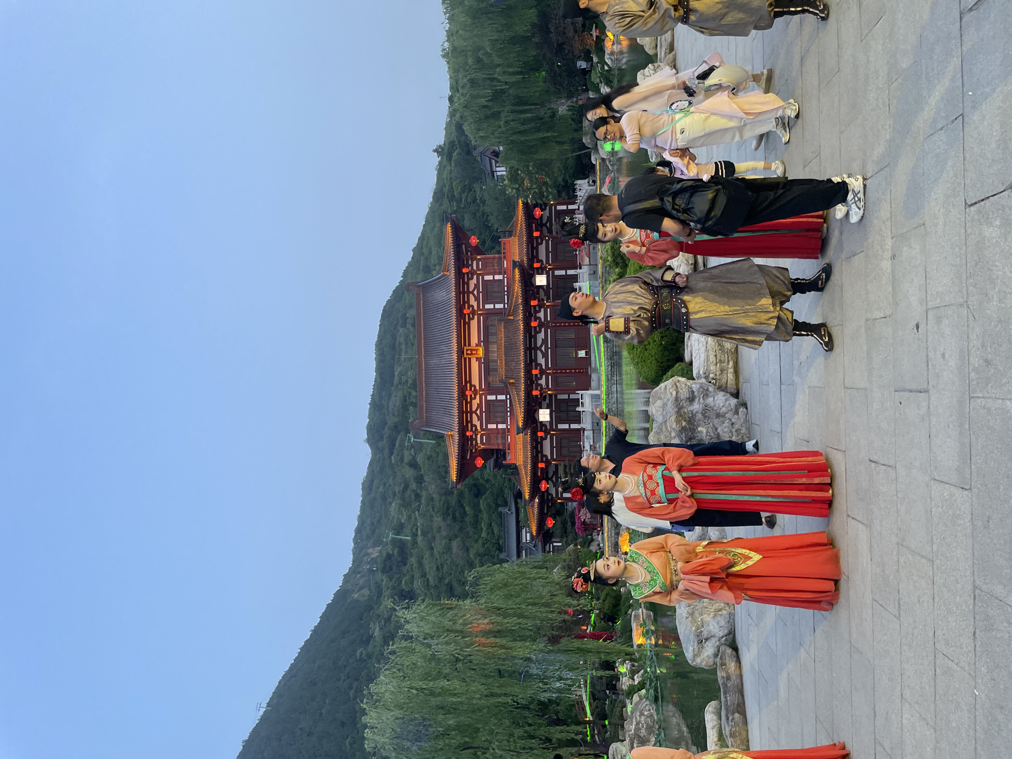In the Lintong District of China, a group of people are positioned on a stone pathway, possibly for a photo opportunity. They appear to be dressed in traditional Chinese attire. Some are looking toward a red and tan building, lit with red lanterns and nestled at the base of a green mountain under a light blue sky. The area is landscaped with rocks and foliage alongside what looks like a pond or stream. Some of the people in the group seem to be taking photos.
