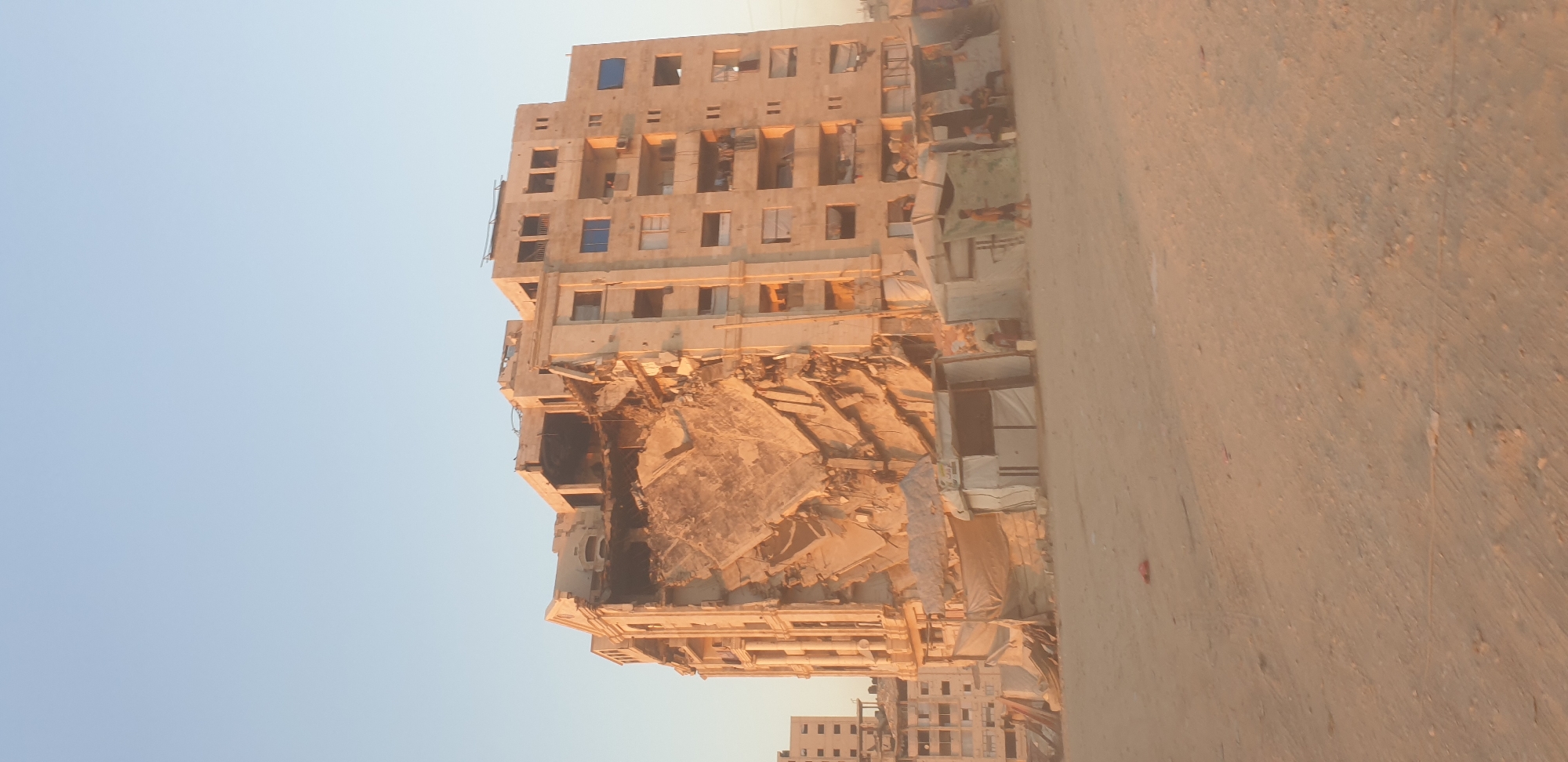 A partially collapsed apartment building stands in Gaza City, Palestinian Territories, under a clear, pale blue sky. The building, a multi-story structure with a beige facade, has sustained significant damage to its left side, with entire sections of walls and floors missing. Debris from the collapse is visible, creating a jagged, open cavity in the building's center. The remaining parts of the building show signs of habitation, with laundry hanging from balconies and some windows intact.
In front of the damaged building, a makeshift settlement of tents and tarpaulins has been erected. Several people are visible among these temporary structures, some sitting and others walking. A few children are present, one walking in the foreground and others interacting near the tents. The ground in front of the settlement and around the building is sandy and appears to be a dirt lot, with scattered debris. In the background, other buildings, some appearing intact and others showing signs of damage, can be seen. The overall scene suggests a community affected by conflict and living in challenging conditions.