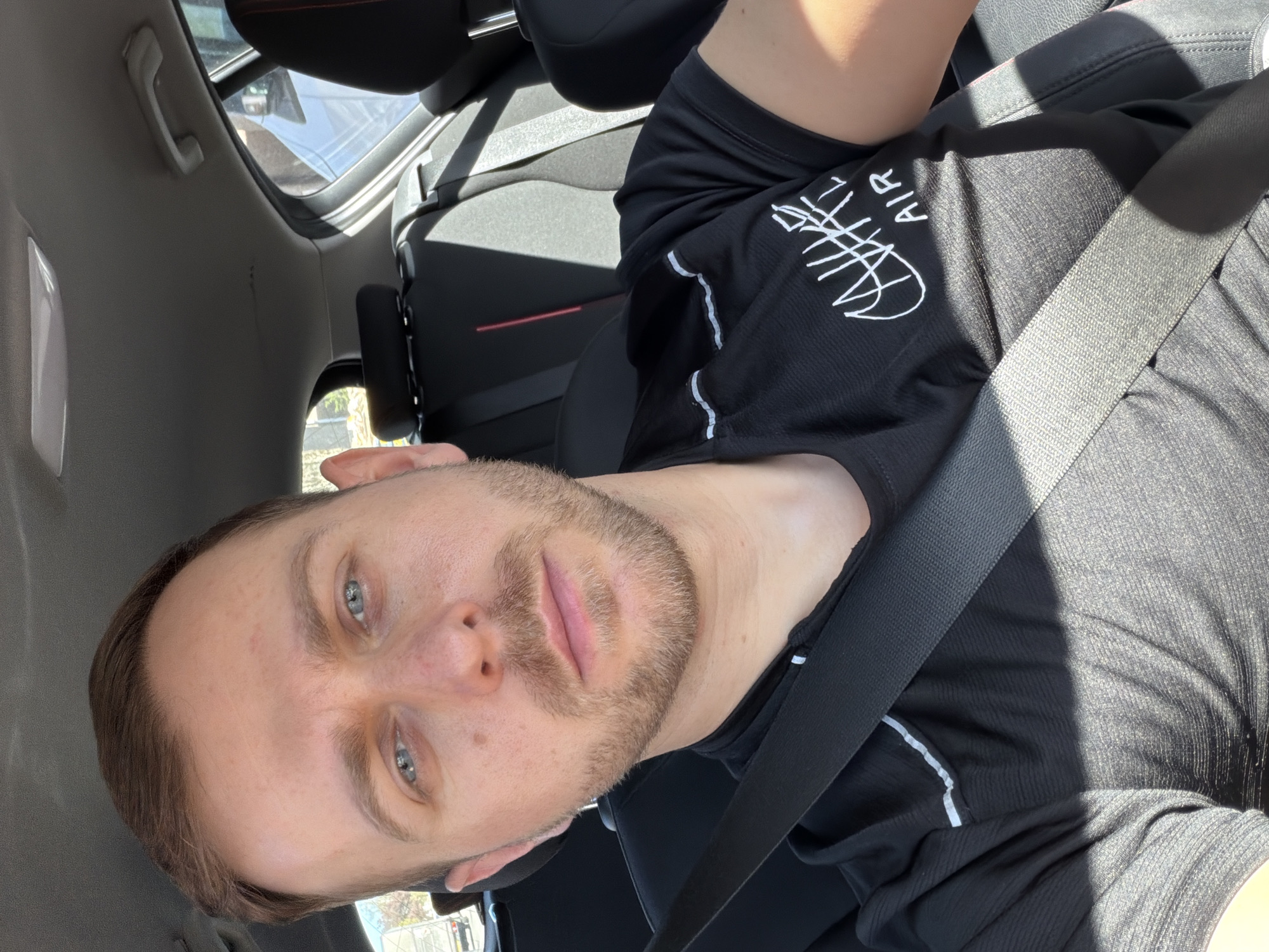 A fair-skinned male with short brown hair and a beard is sitting in a car, wearing a black shirt with a white logo. He is wearing a seatbelt. The interior of the car is gray and black. The location is unknown.