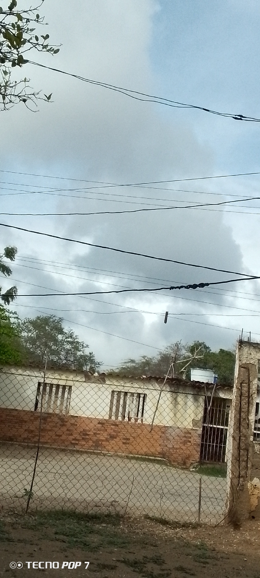 The image depicts an overcast sky with numerous power lines crisscrossing the frame. In the foreground, a chain-link fence encloses a yard with sparse vegetation and some dirt. Beyond the fence, a low-lying building with a tiled roof is visible. The building has two windows with bars and appears to be constructed of light-colored stucco over brick. Behind the building, trees and foliage are present, and a white water tank is visible on the roof. To the right, a section of a partially collapsed concrete structure stands. The scene appears to be a residential area. The bottom left corner of the image displays the watermark "TECNO POP 7". This scene is consistent with the typical appearance of residential areas in Altagracia, Venezuela, characterized by basic construction, the prevalence of power lines, and the presence of vegetation.