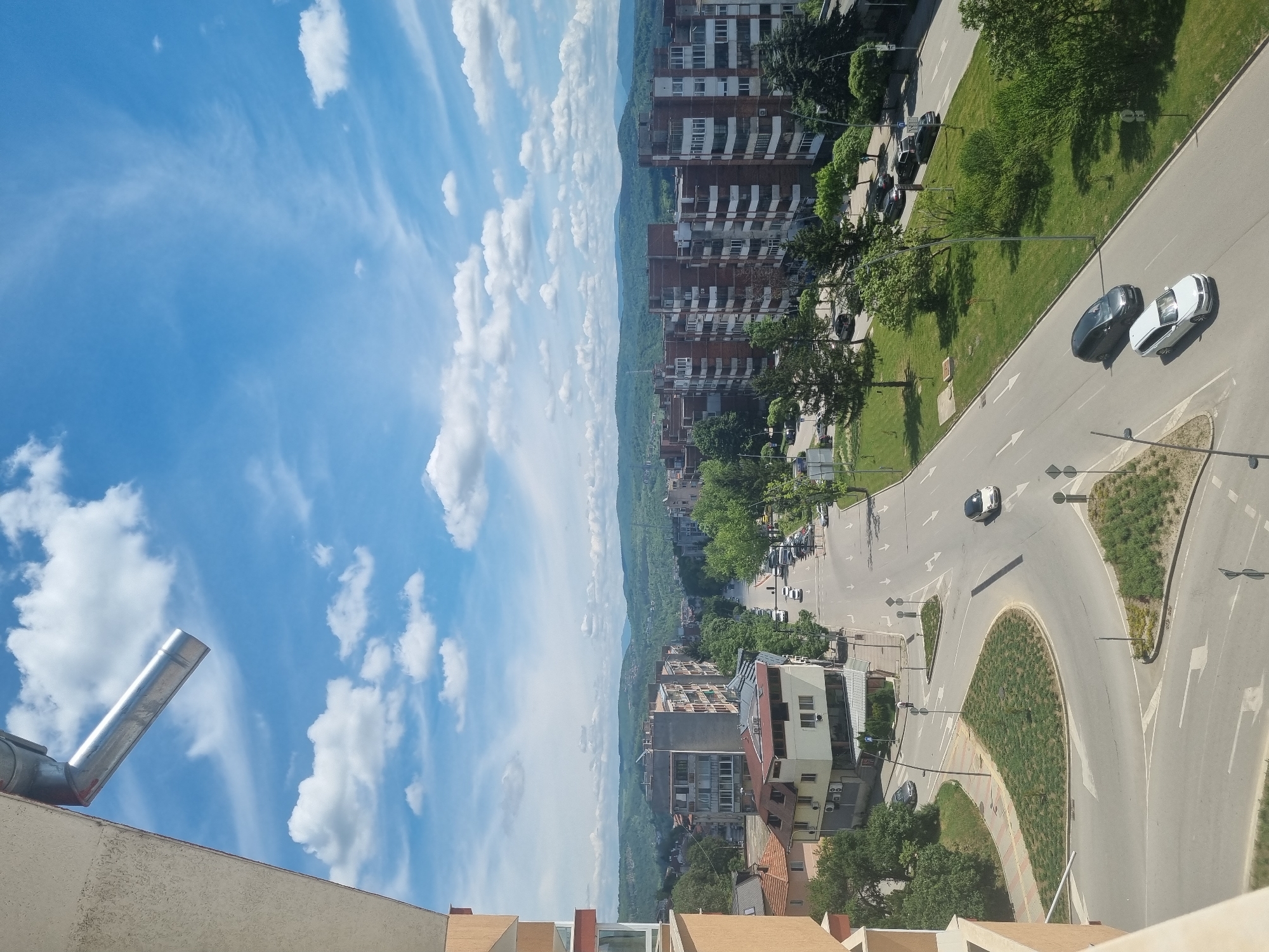 The image shows an elevated view of a street scene in Veliko Tarnovo, Bulgaria. The sky is blue with scattered white clouds. In the distance, a range of green hills is visible.
The foreground consists of a rooftop with a metal chimney. The street below has a roundabout with landscaping in the center. Cars are driving on the road. Several multi-story brick apartment buildings are visible along the street, interspersed with green spaces and trees. A roundabout and roads are visible. There are several cars in the street.