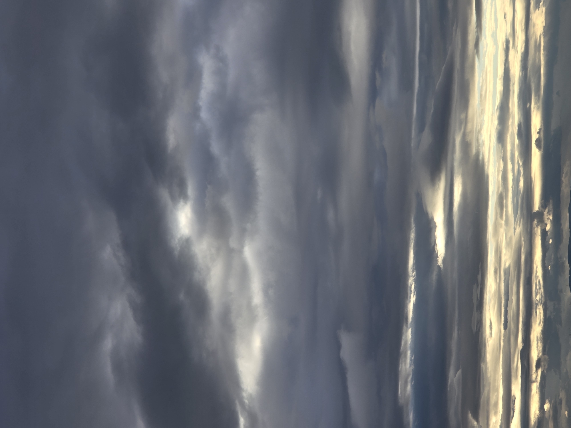 The image shows a cloudy sky with varying shades of gray and white. Some clouds are dark and dense, while others are lighter and more translucent, with a hint of golden light filtering through them. The clouds appear to be vertical in orientation, possibly due to a strong wind or atmospheric conditions. The specific location is unknown.