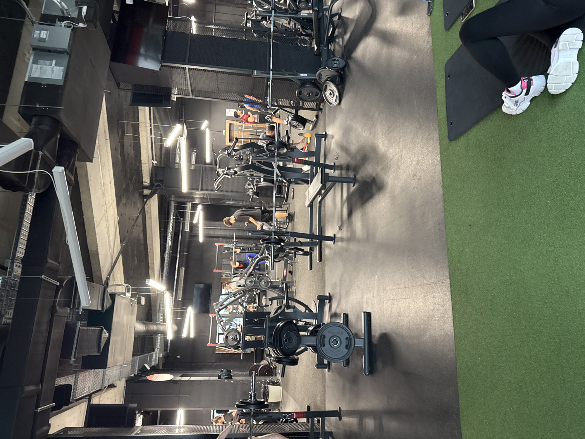 The image shows the interior of a gym. Several weightlifting machines are visible, and individuals are using the equipment. The floor is made of both black rubber and green artificial turf. A person wearing black leggings and white sneakers is partially visible in the upper right corner of the frame. The gym features industrial-style architecture with exposed ventilation systems and lighting fixtures. The location in Plovdiv, Bulgaria, is not discernible from the image.