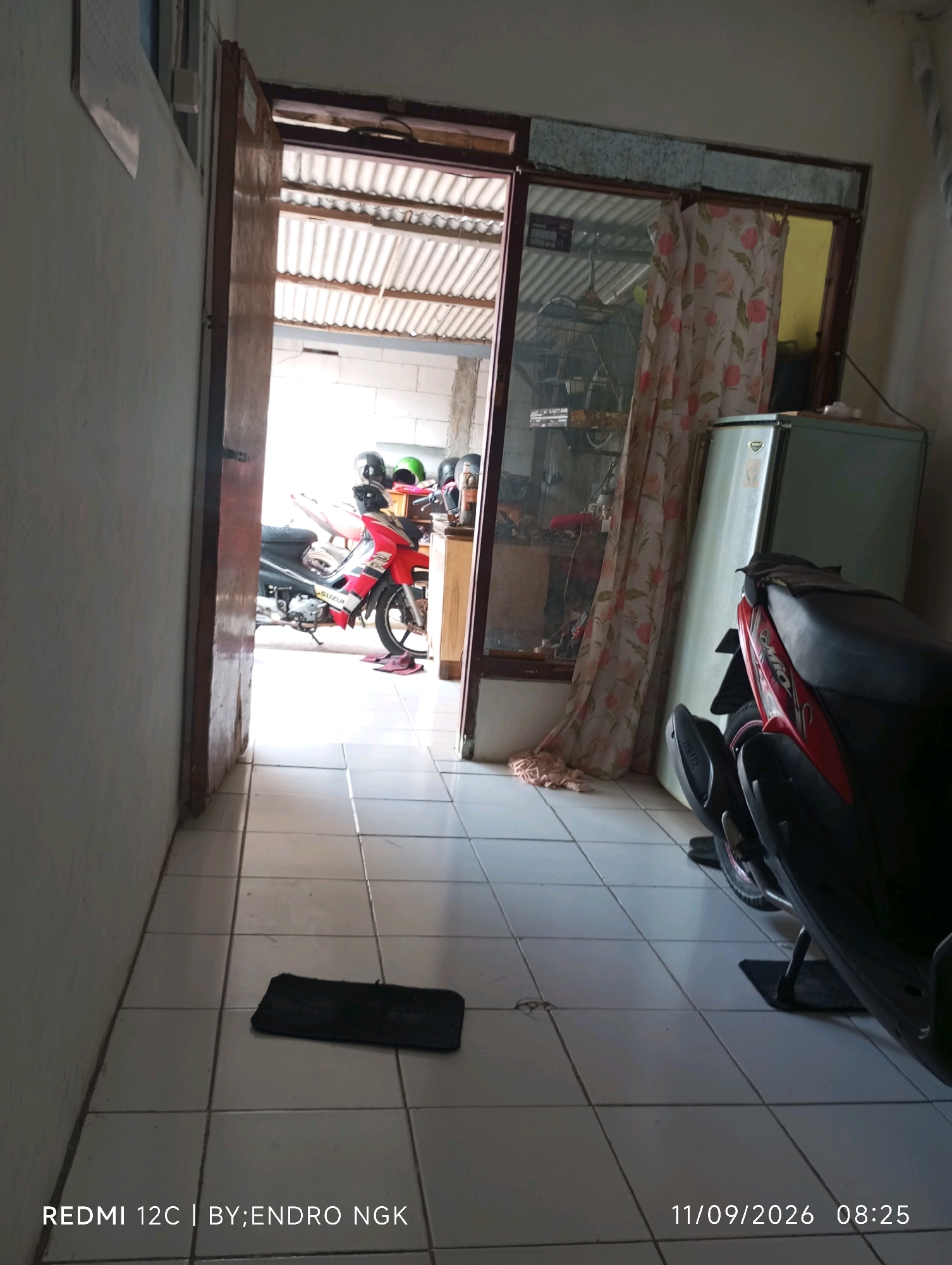The image shows a view of an interior space, possibly a garage or a covered outdoor area, with a doorway leading to the outside. In the foreground, a tiled floor is visible, with a black rectangular mat lying near the center. To the right, the back half of a red and black motorcycle is parked. Further into the space, through an open doorway, several motorcycles are parked, along with various helmets. A light green refrigerator stands near the right edge of the frame, partially obscured by the motorcycle. A floral patterned curtain hangs near the refrigerator. The overall impression is of a storage area for motorcycles. The text at the bottom of the image indicates that the photo was taken with a Redmi 12C by Endro NGK on 11/09/2026 at 08:25. While the specific location in Ringinanom, Indonesia is not visually discernible from the image itself, the text implies it was taken there.