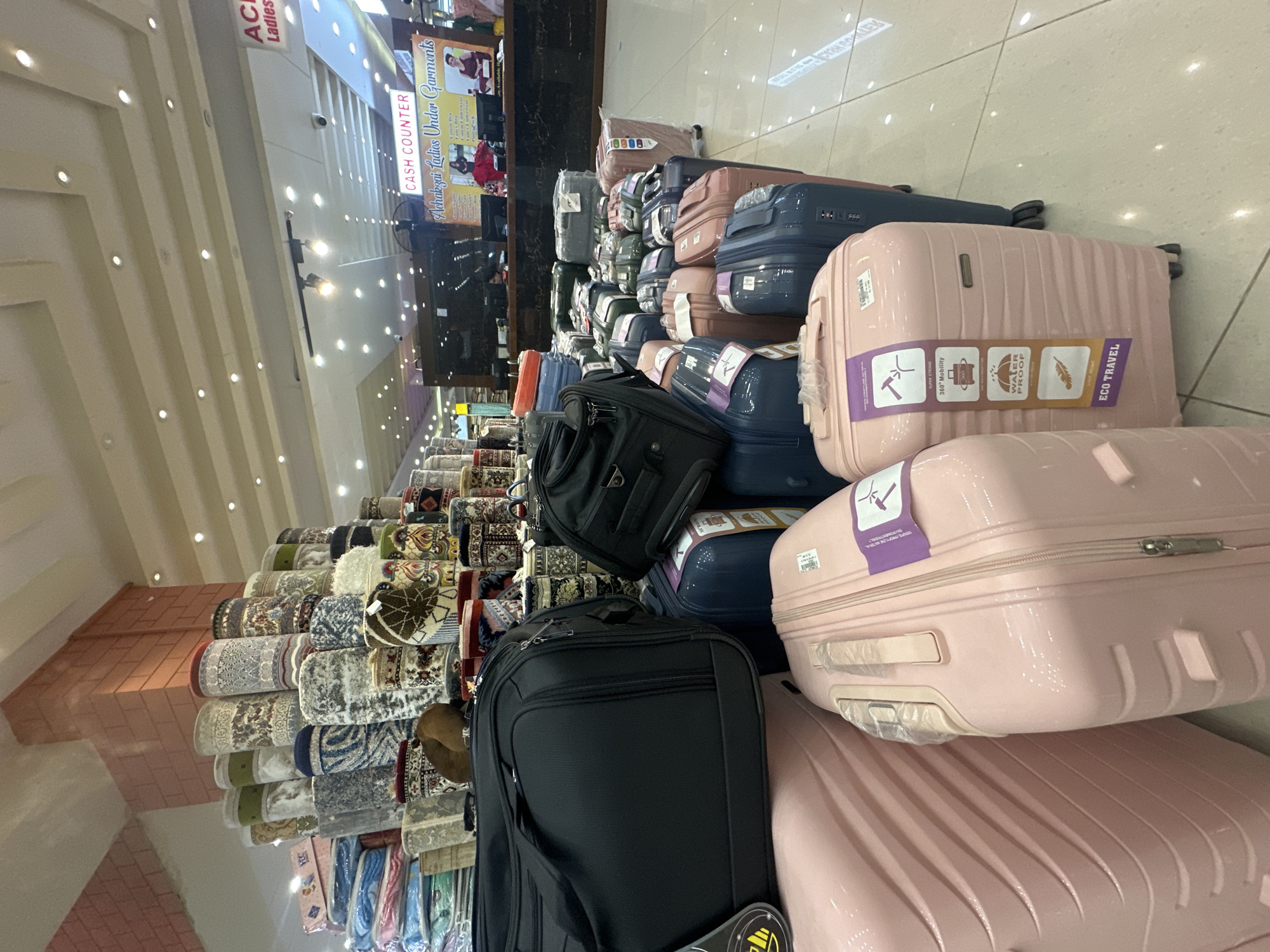 The image depicts an interior shot inside what appears to be a retail store. The location appears to be Achakzai Ladies Under Garments in Quetta City Tehsil, Pakistan, based on the signage present. Stacks of rolled carpets and rugs are visible on the left side of the frame. Toward the center and right, there are multiple suitcases, including pink and blue ones, with some having informational stickers indicating features like "360° Mobility," "Water Proof," and "ECO Travel." The suitcases are arranged in a somewhat haphazard manner, suggesting a display of merchandise. In the background, above the carpets, there is a "CASH COUNTER" sign. The overall environment seems to be brightly lit with numerous ceiling lights.