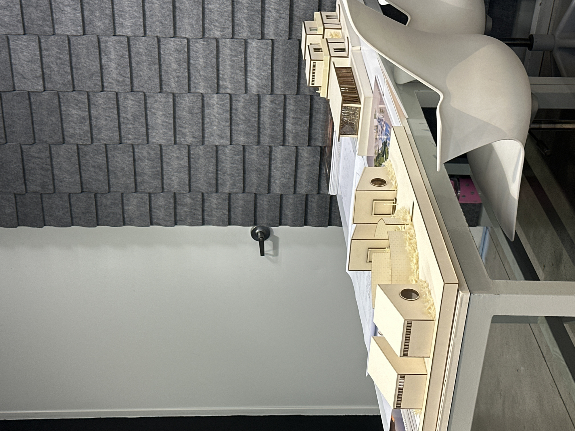 The image shows a gray textured wall covering the top of the frame. Below the wall is a plain white wall, and near the bottom is a black floor. A small dark handle protrudes from the white wall. A table with several model buildings sits at an angle. The model buildings are cream-colored and have small details. Next to the models is a white chair. The scene is inside an office. The location is unknown.