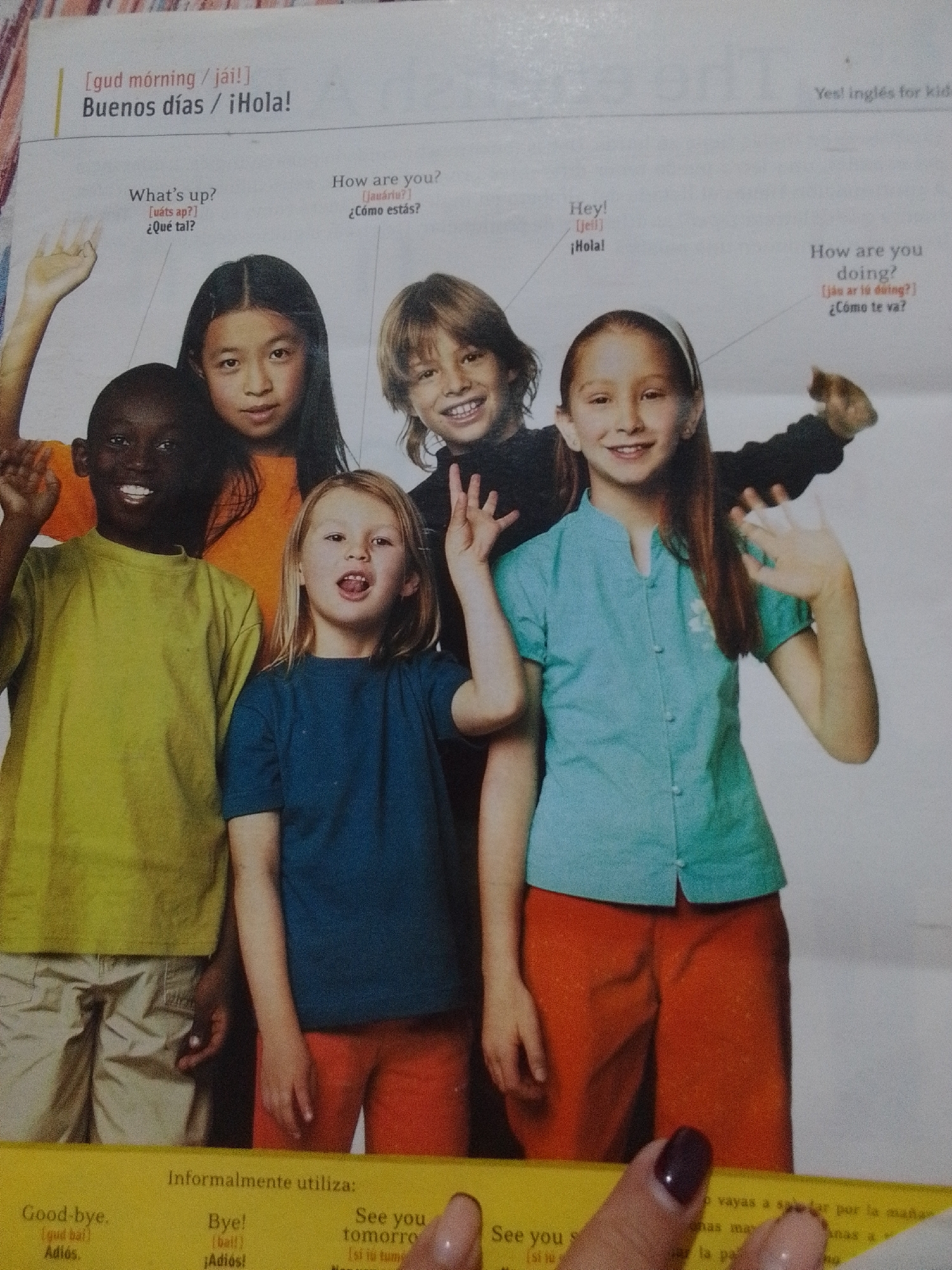 This is a close-up, high-angle shot of a page from an English language learning book or workbook, featuring a photograph of five young children posing and waving. The children are of diverse ethnicities and are smiling. Text overlays the photograph, providing greetings in English and Spanish. The background is a plain, light color. The bottom of the page contains additional text in a yellow border. There is no visible indication of La Tagua, Venezuela within the image.