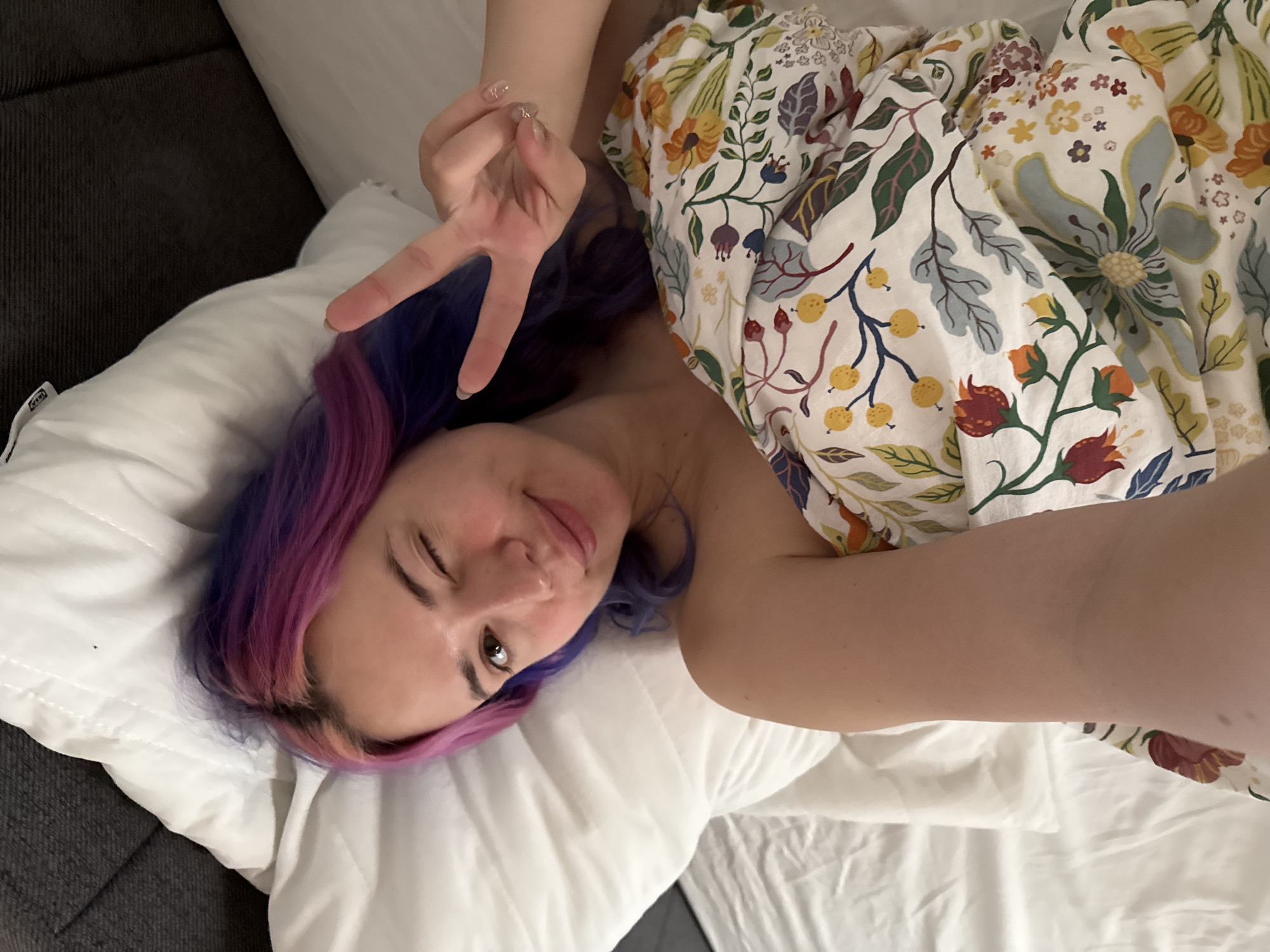 A young woman with distinctive multi-toned hair, featuring shades of purple and pink/magenta, is captured in a relaxed indoor setting, likely in the City of Zagreb, Croatia. She is lying down, possibly on a bed or a sofa, and gazes directly towards the camera with a playful expression, winking one eye and offering a gentle smile. Her right hand is raised to form a peace sign, revealing manicured nails with a subtle glitter finish. Her head rests comfortably on a soft white pillow, which bears a small, visible "IKEA" tag, indicating a potential furnishing source for the location. A vibrant, white-based duvet or blanket, adorned with an intricate pattern of colorful flowers, leaves, and berries in orange, yellow, green, and blue hues, covers part of her body. A dark grey upholstered surface, possibly a bed frame or another piece of furniture, is partially visible on the left side of the frame. The lighting is soft, contributing to the casual and intimate atmosphere of the photo, which conveys a sense of cheerfulness and ease.