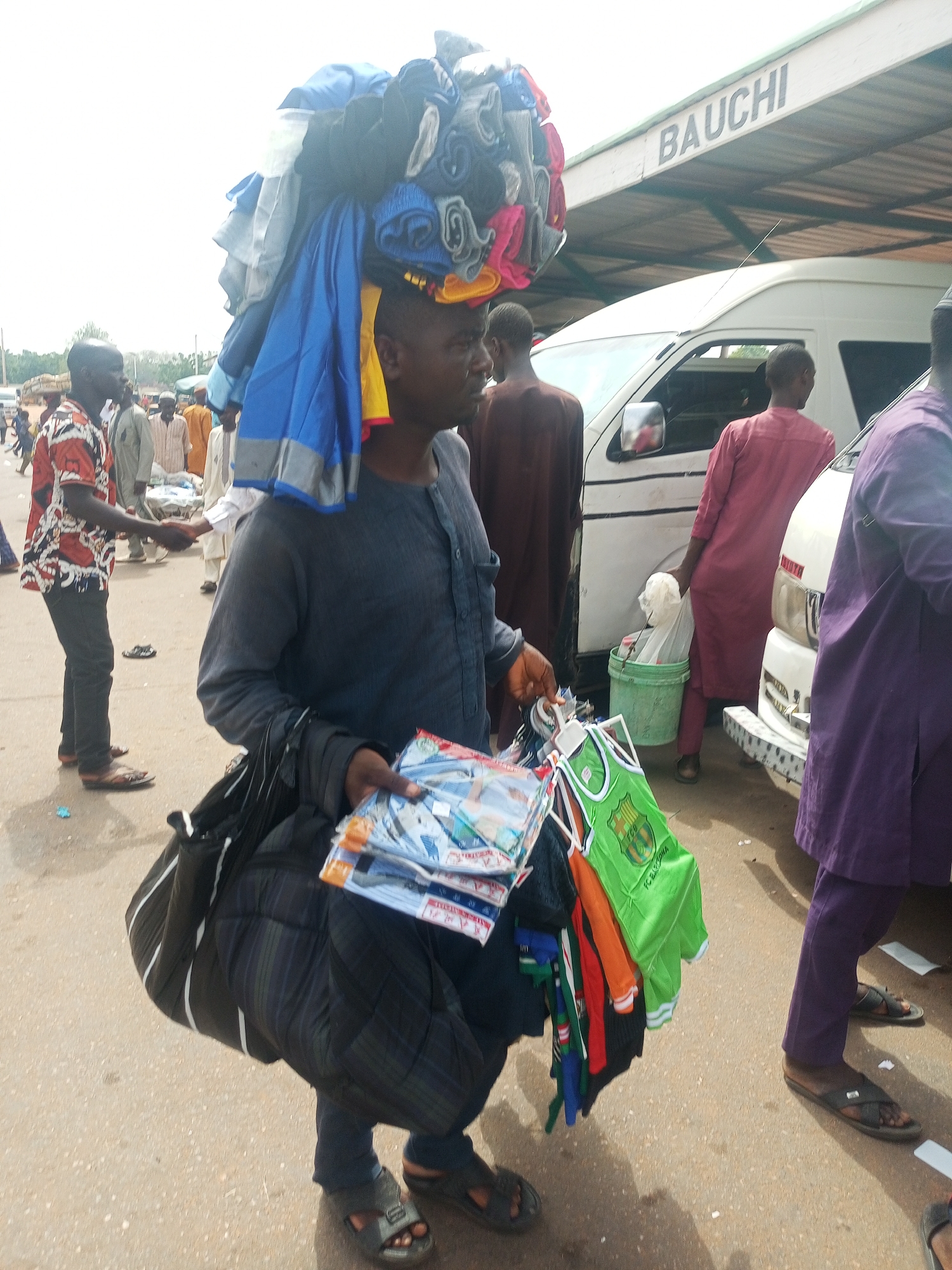 This daytime image captures a bustling street scene, likely in Gombe, Nigeria, depicting a street vendor as its central subject. The man, dressed in a dark long-sleeved top, dark plaid trousers, and sandals, carries a substantial bundle of clothes on his head. In his left hand, he holds several packaged garments, including what appears to be underwear or socks, alongside various small items of clothing, notably a bright green FC Barcelona football jersey. He also carries a black shoulder bag slung over his left arm.
The background reveals a lively market or transport hub. Several other men are present; one to the left wears a patterned shirt and dark trousers, while others are seen standing, walking, or engaging in conversation. On the right side of the frame, a white commercial passenger van is parked, and a man in a reddish-brown outfit is visible next to it, seemingly attending to a green bucket. Another individual in a purple garment is partially visible on the far right.
The ground is a reddish-brown, dusty surface, suggesting an unpaved or lightly paved outdoor area. The lighting is bright and even, indicating either an overcast day or the middle hours of the day. A prominent sign bearing the word "BAUCHI" is visible on a roof structure above the white van, likely denoting a destination for the transport vehicles or a local landmark. The scene vividly portrays the dynamic daily life and informal commerce typical of a busy urban or semi-urban environment in Nigeria.