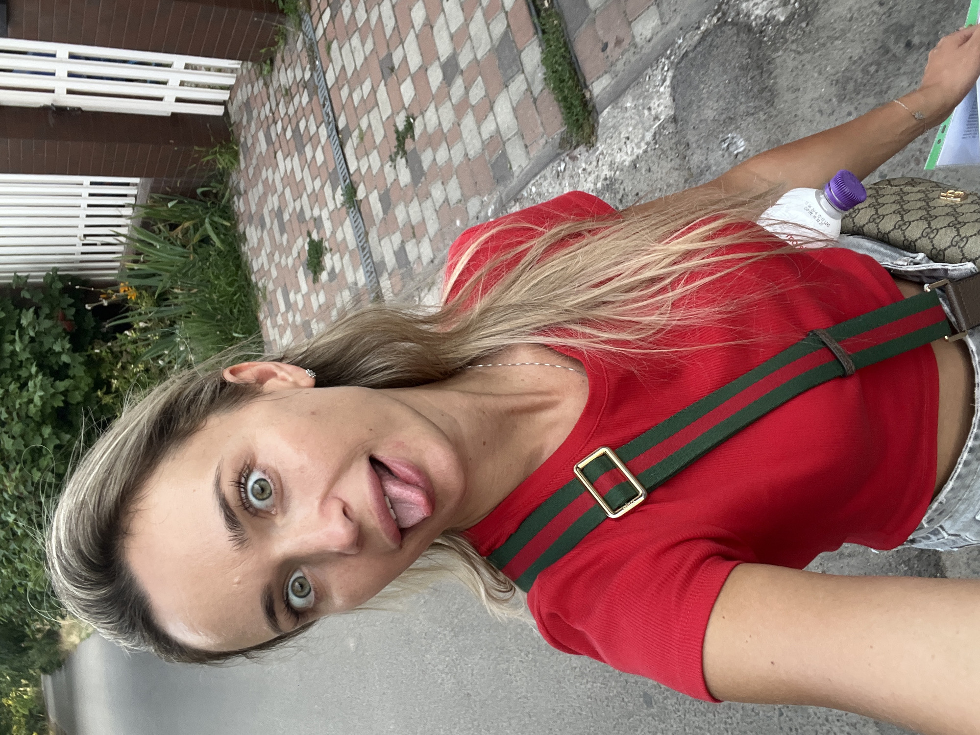A young Caucasian woman with blonde hair and green eyes stands on a sidewalk with a paved path to her left and a brick-tiled walkway to her right, leading to a white wooden fence and a brown brick building. She is wearing a red t-shirt and denim shorts, with a green and red striped strap across her chest. A patterned crossbody bag is slung over her shoulder. The woman is sticking out her tongue and making a playful face at the camera. A bottle of water with a purple cap is tucked into her waistband. The environment appears to be outdoors, possibly in a residential area.