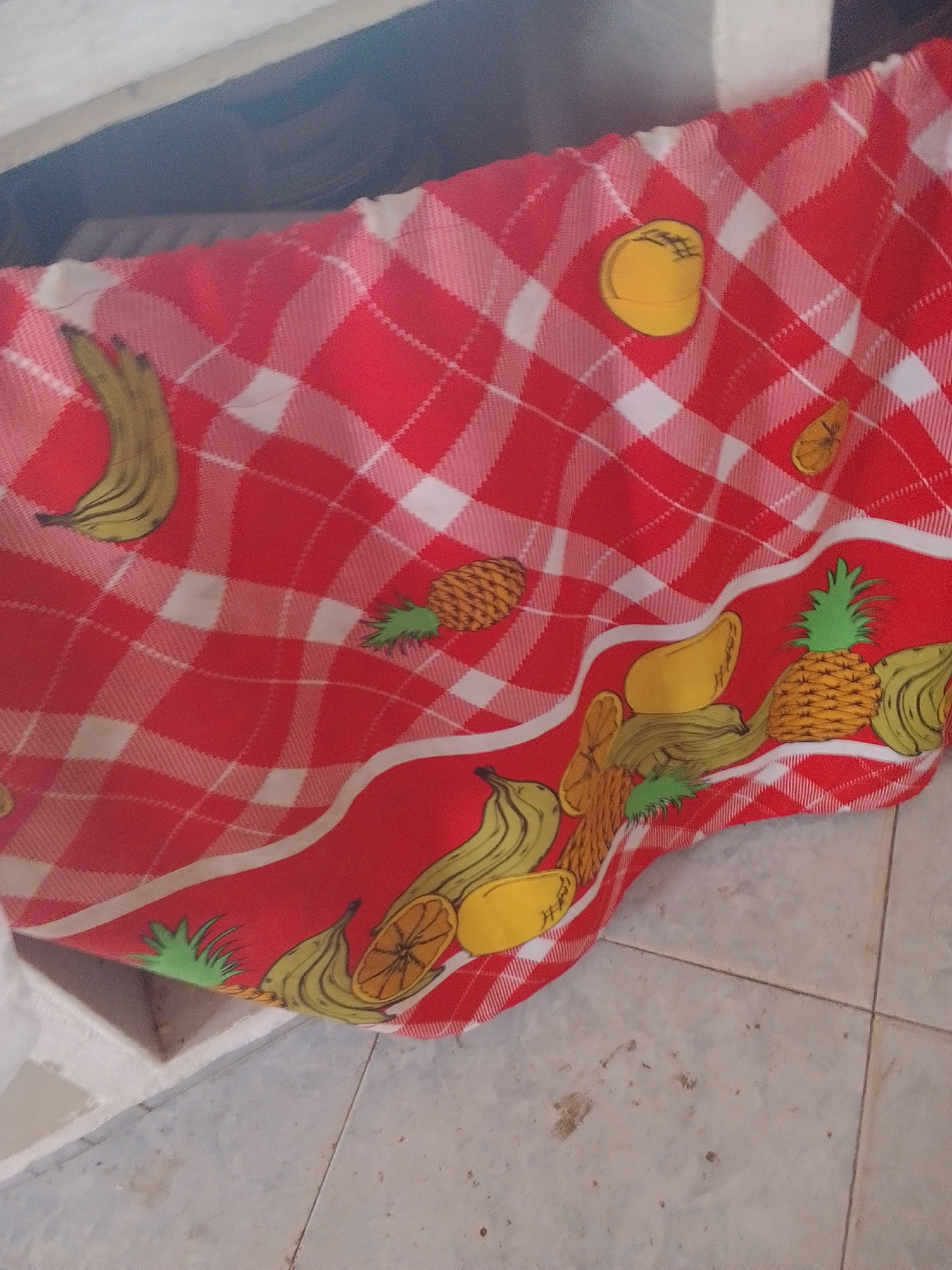 The image shows a close-up of a red and white checkered fabric with a fruit pattern. The fruits depicted include bananas, pineapples, oranges, and mangoes. Some of the fruits are whole, while others are shown as slices. The fabric appears to be draped over the edge of a white surface, possibly a table or shelf, and part of a tiled floor is visible below. The lighting suggests it is daytime. There is no text visible in the image. The context provided is La Tagua, Venezuela.