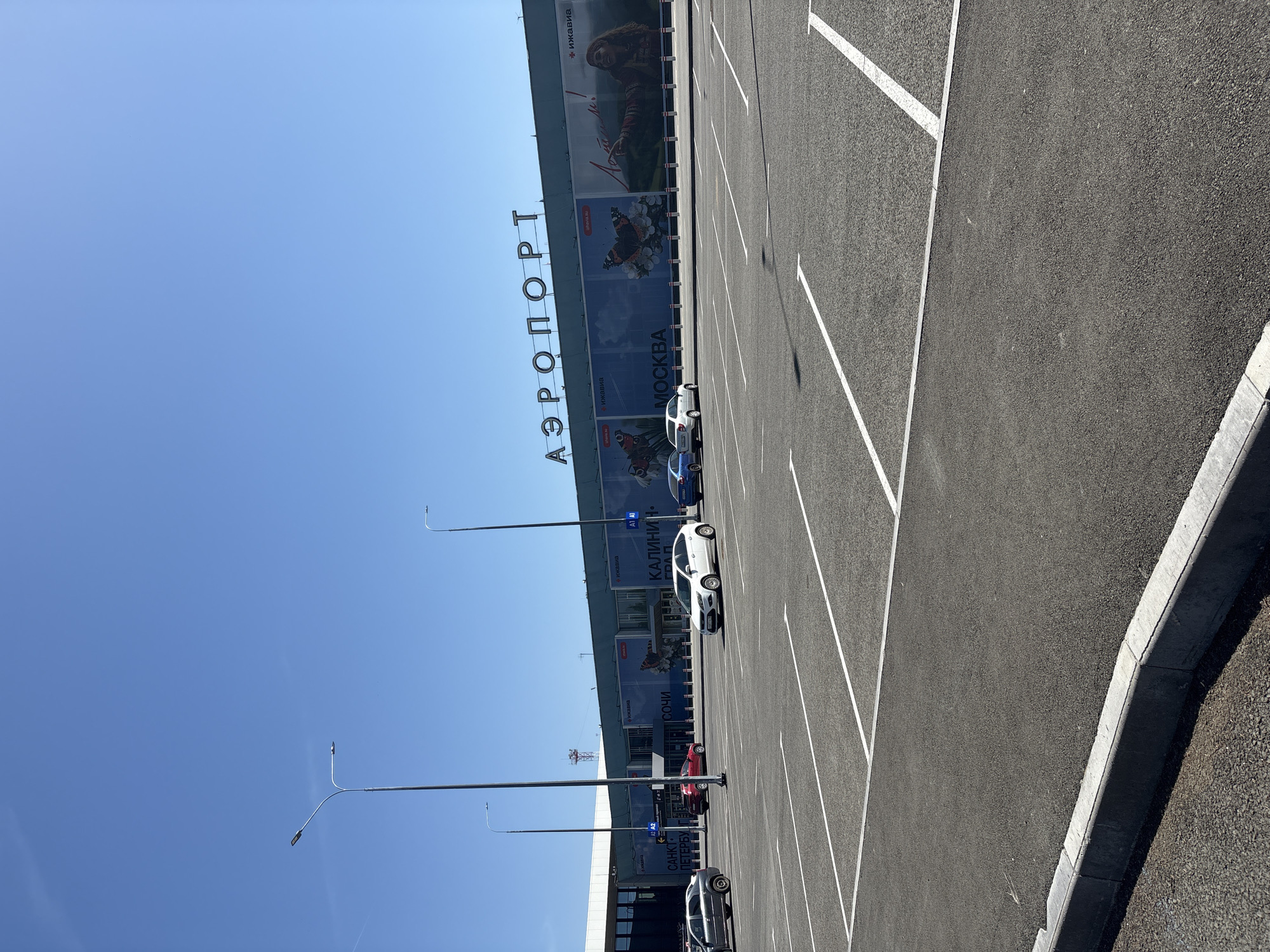 The image shows a parking lot at an airport with a large sign that says "AЭРОПОРТ MOCKBA". There are several cars parked in the lot. The parking lot and airport are likely located in or near the city of Moscow, Russia. The text includes the word "ижавиа", that is likely the name of an air company.
There are also posters visible with the locations of other cities like, "CANKT, ПЕТЕРБУ, СОЧИ".